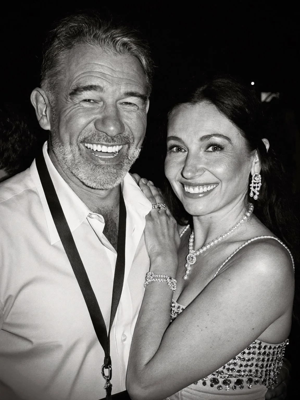 A smiling older man with gray hair and beard wearing a white shirt and a lanyard, and a woman with dark hair, jewelry, and a patterned dress, both smiling for a photo in black and white.