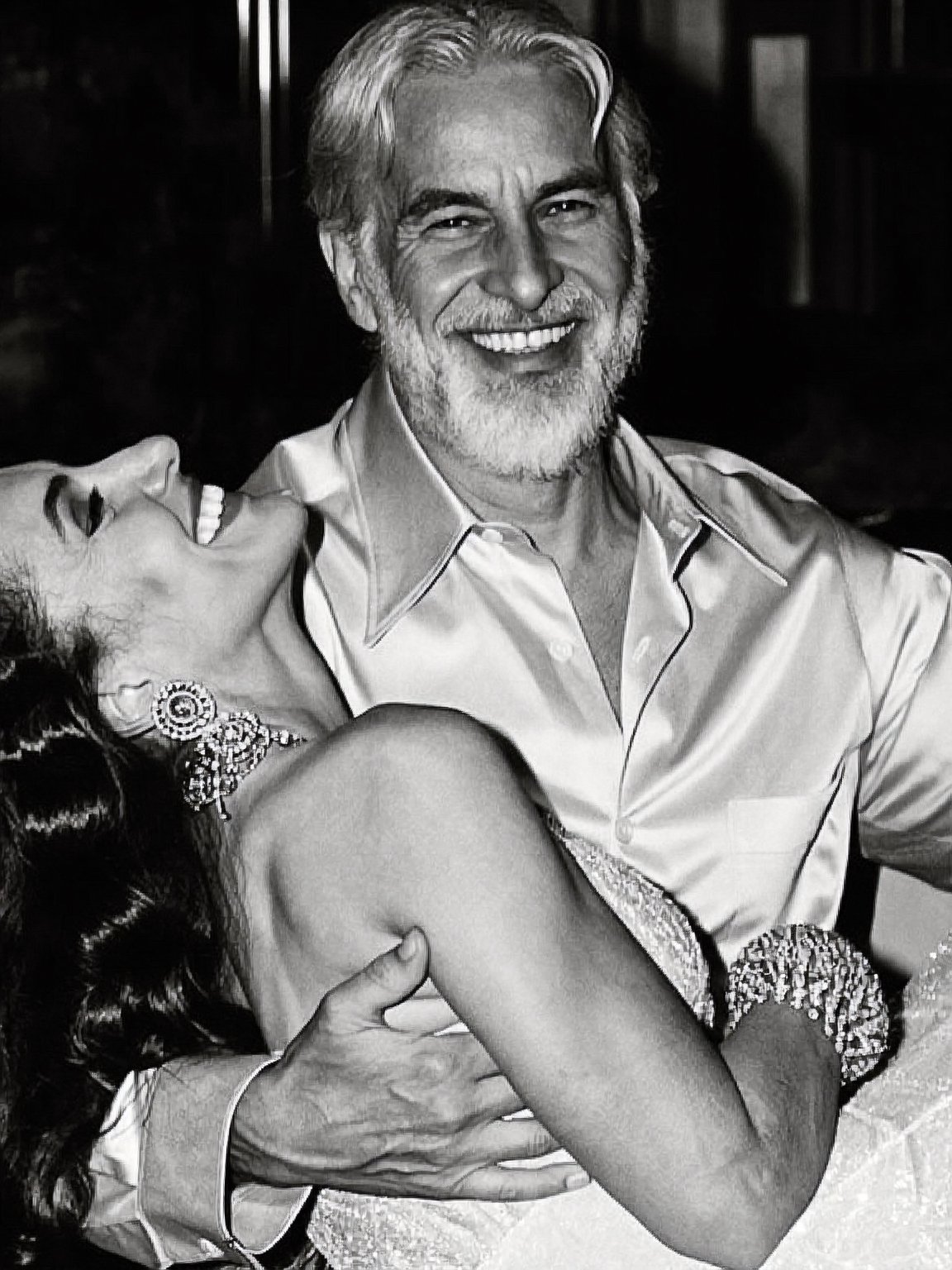 A smiling man with a beard and mustache wearing a button-up shirt, being embraced and kissed on the cheek by a woman in a dress with earrings.