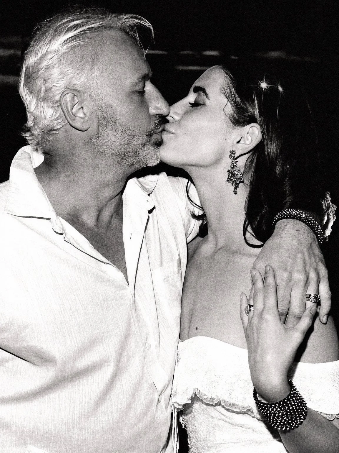 A black and white photo of an older man and a woman sharing a kiss, with their eyes closed. The man has gray hair and a beard, wearing a light-colored shirt. The woman has dark hair, earrings, and is wearing a strapless dress with jewelry.