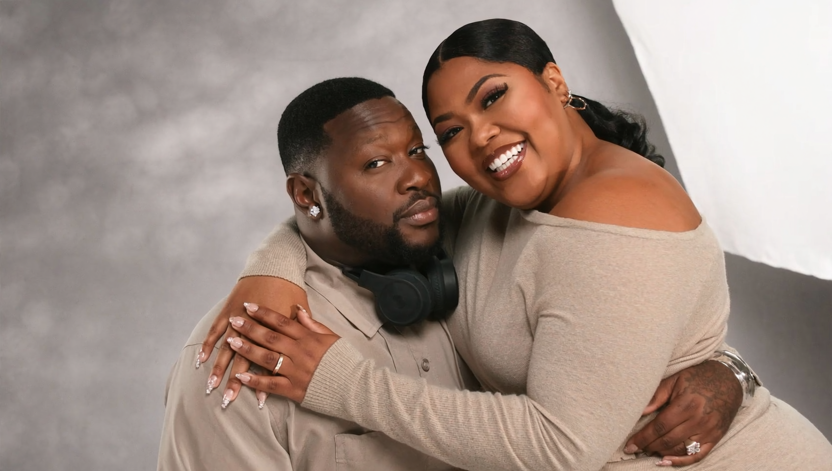 A couple hugging, smiling, and looking at the camera against a neutral background. The woman has long dark hair, a beige off-the-shoulder top, and jewelry. The man has short dark hair, a beard, earrings, and headphones around his neck.