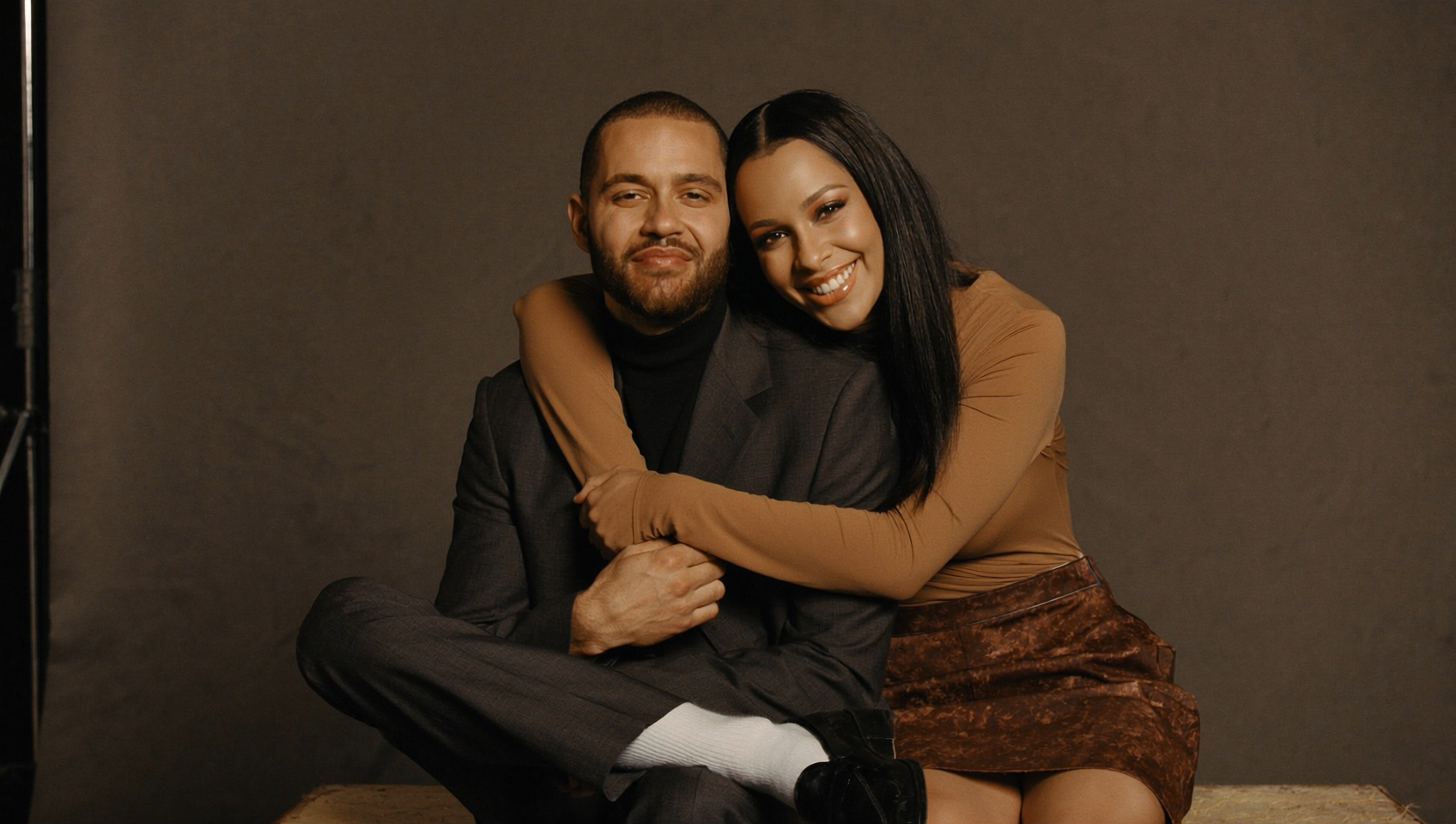 A man and woman sitting together, smiling, with the woman hugging the man from behind on a plain dark background.