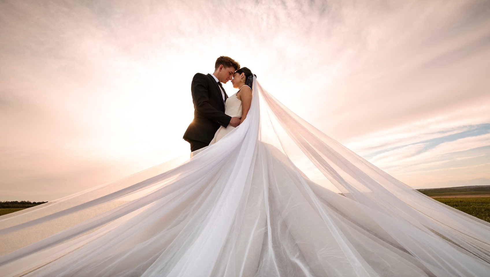 A newlywed couple standing close together at sunset in an open field, the bride wearing a long white wedding gown with a train, and the groom in a black suit, with their foreheads touching.