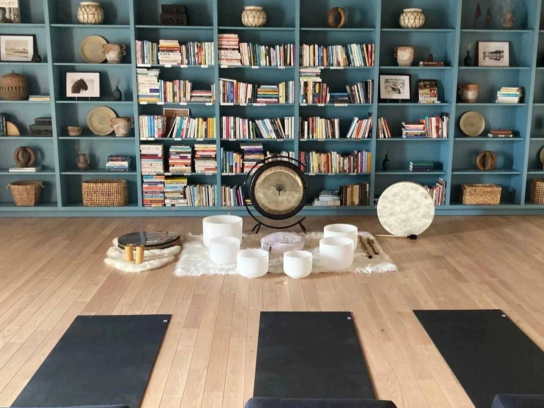 Yoga mats on wooden floor facing a shelf filled with books and decorative objects, with crystal singing bowls and a gong in front.