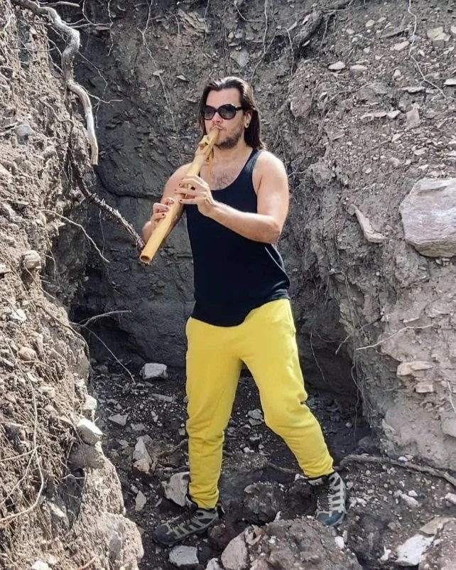 A person with long hair, wearing sunglasses, a black tank top, yellow pants, and hiking shoes, stands in a rocky outdoor setting and plays a wooden flute.