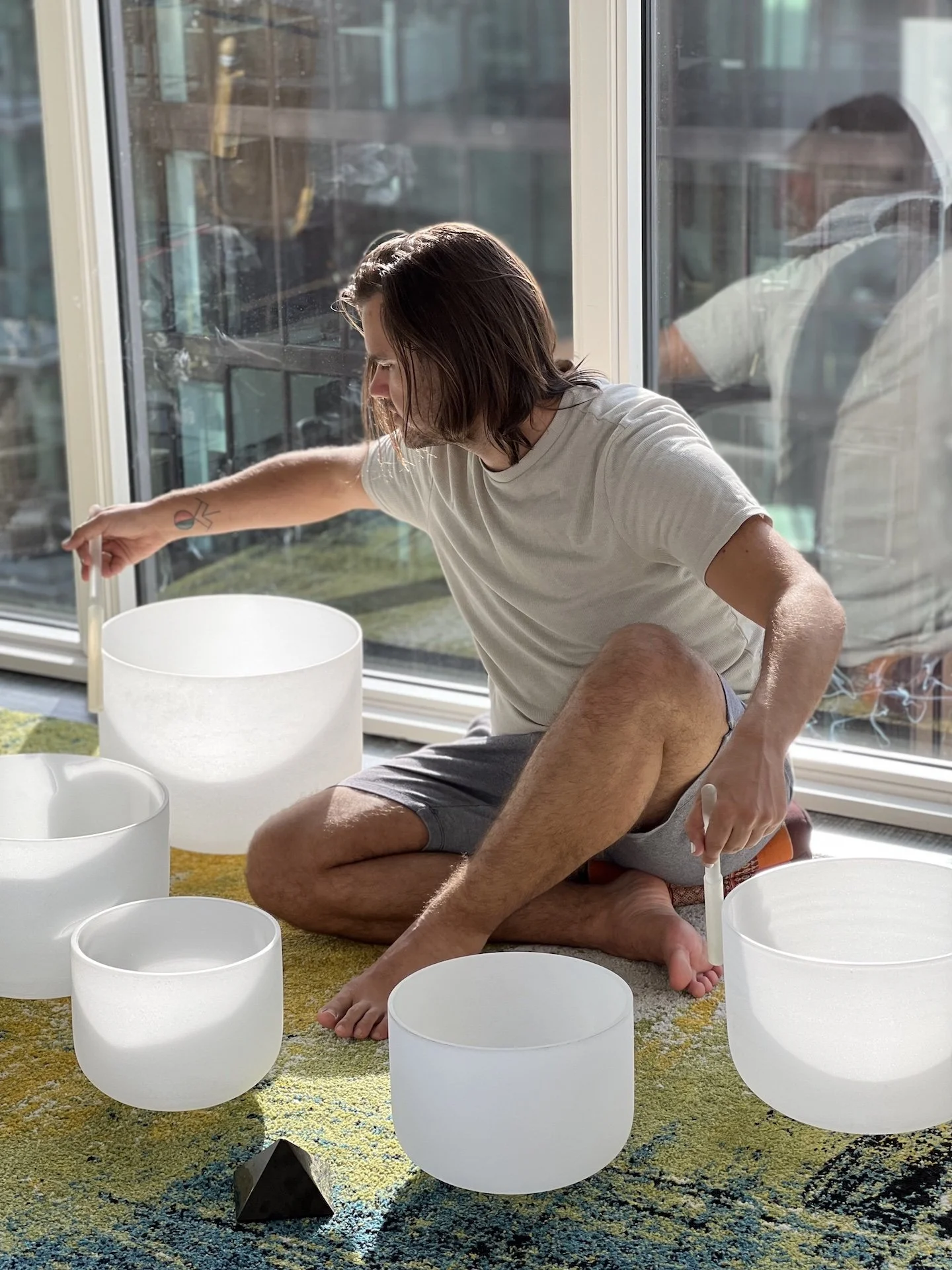 A person with shoulder-length hair playing crystal singing bowls on a colorful carpet near a large window in a sunlit room.