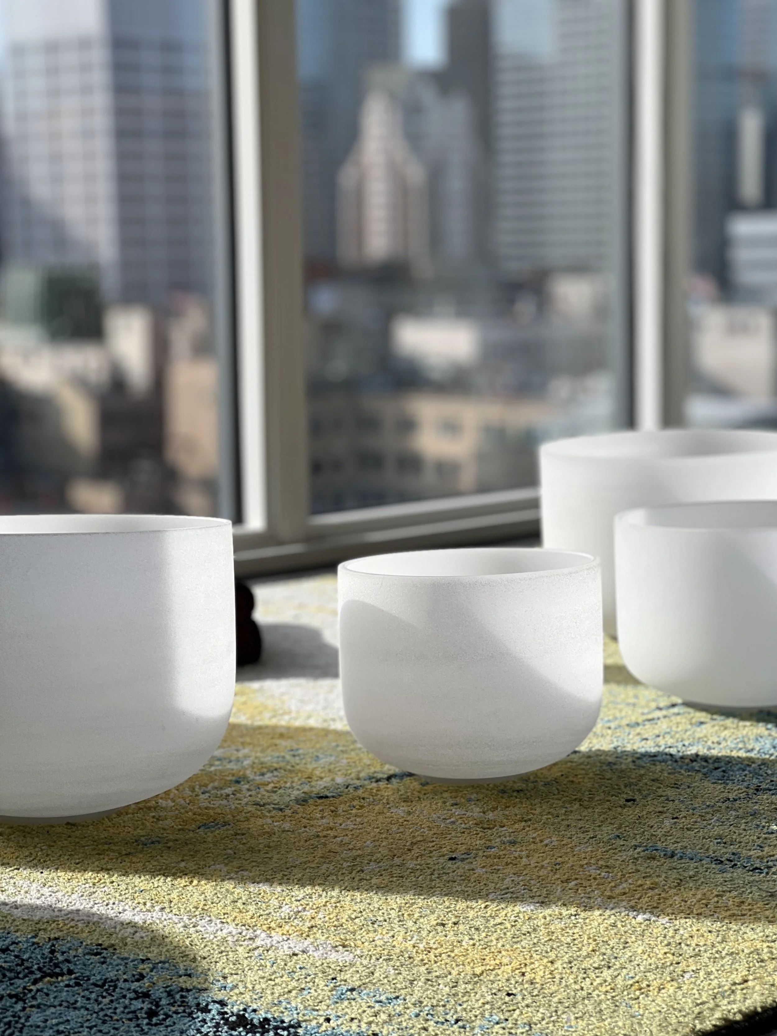 Four white crystal singing bowls on a colorful rug with a Los Angeles skyline view through large windows.