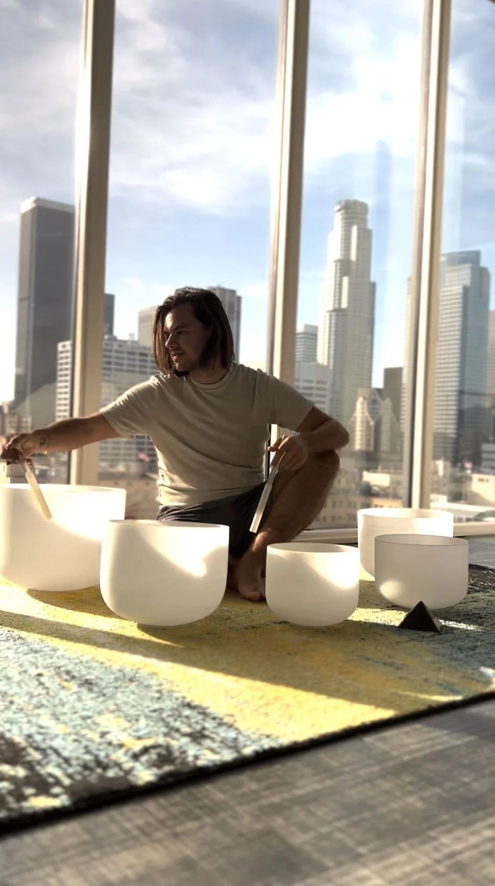 A person with long hair playing crystal singing bowls in a high-rise building with a city skyline in the background.