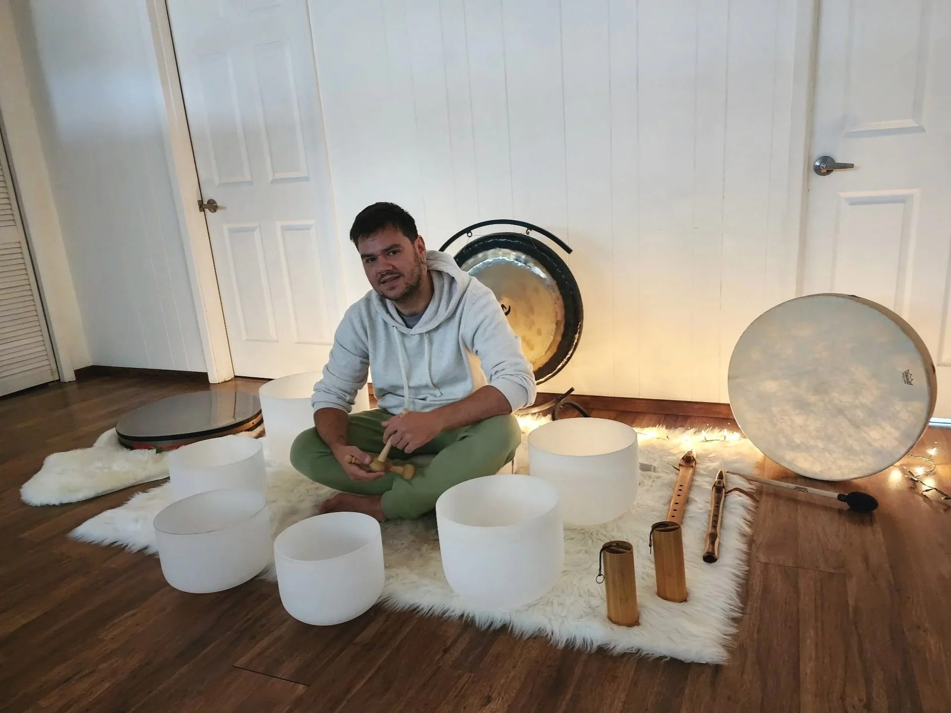 A person sitting cross-legged on a white fur rug, surrounded by singing bowls, wooden flutes, and a gong, with a white wall and doors in the background.