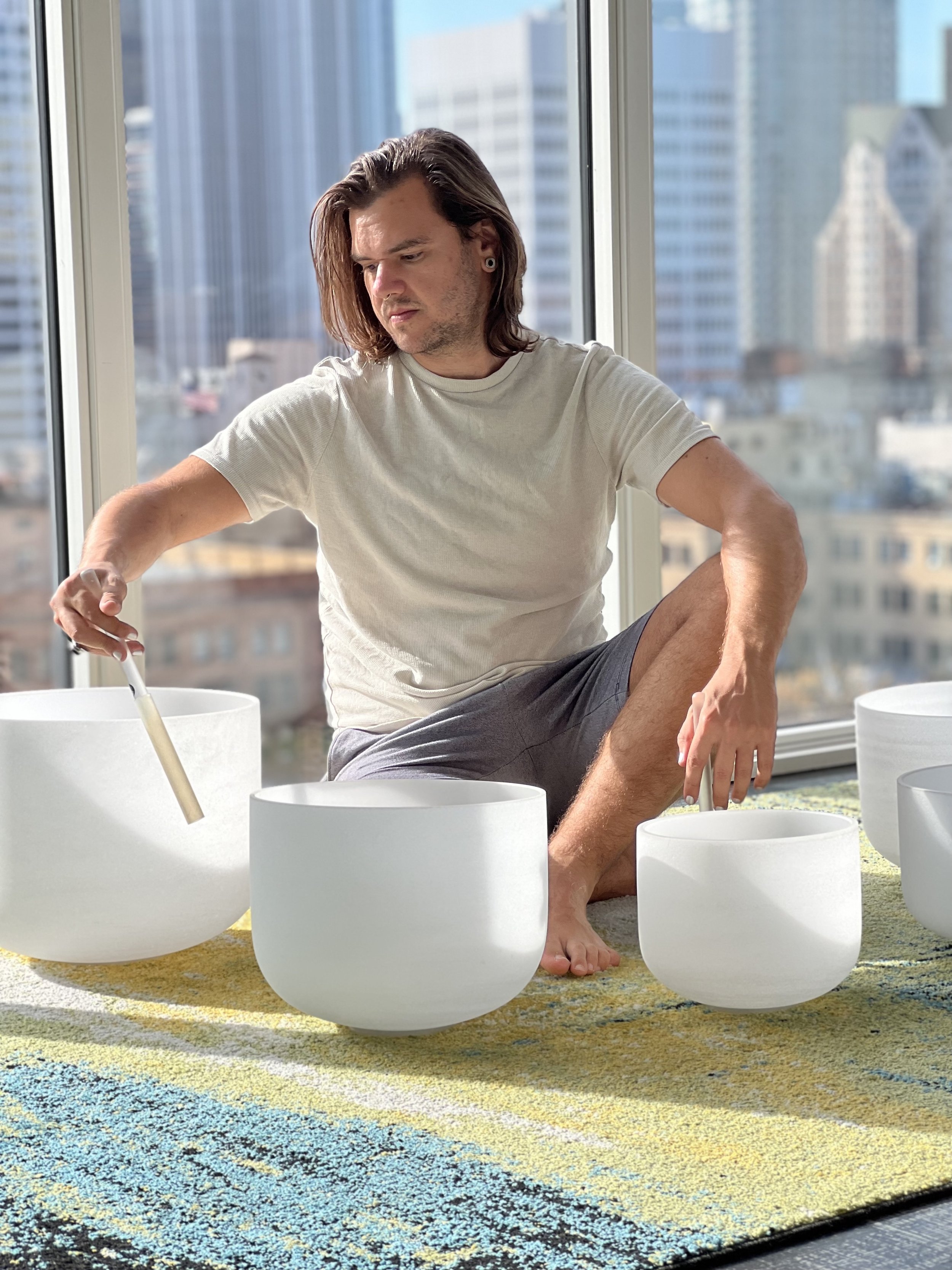 A person sitting on the floor playing crystal singing bowls by a large window in a city apartment with tall buildings in the background.