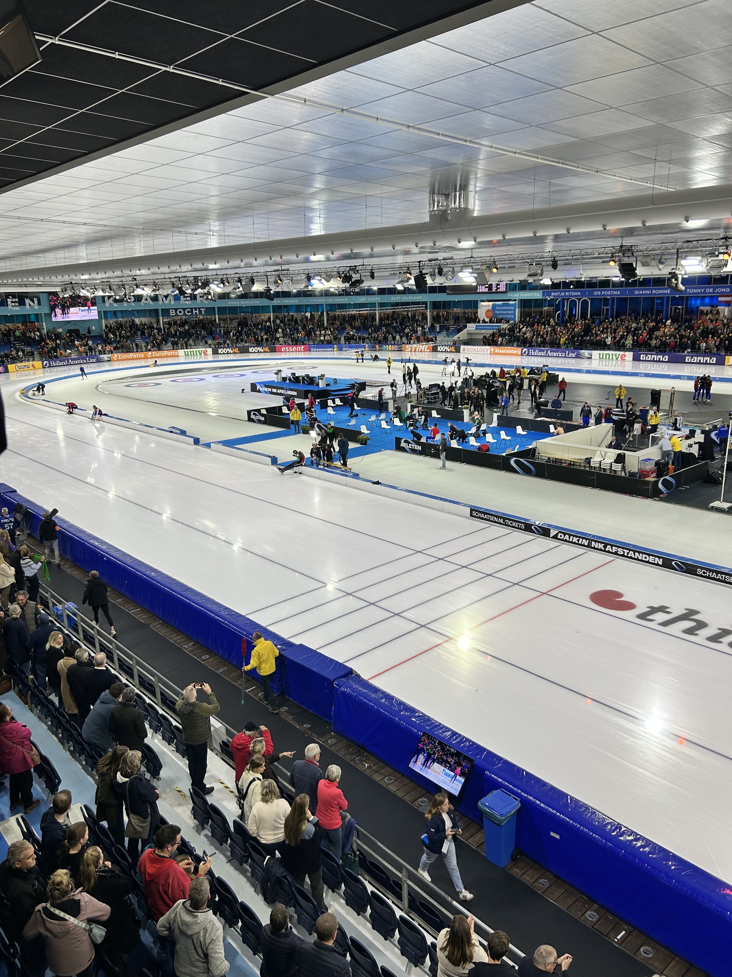An indoor ice skating rink with spectators seated and standing along the sides, some watching and some taking photos. The ice surface is prepared with markings and the arena is well-lit with overhead lights and a large crowd in the background.