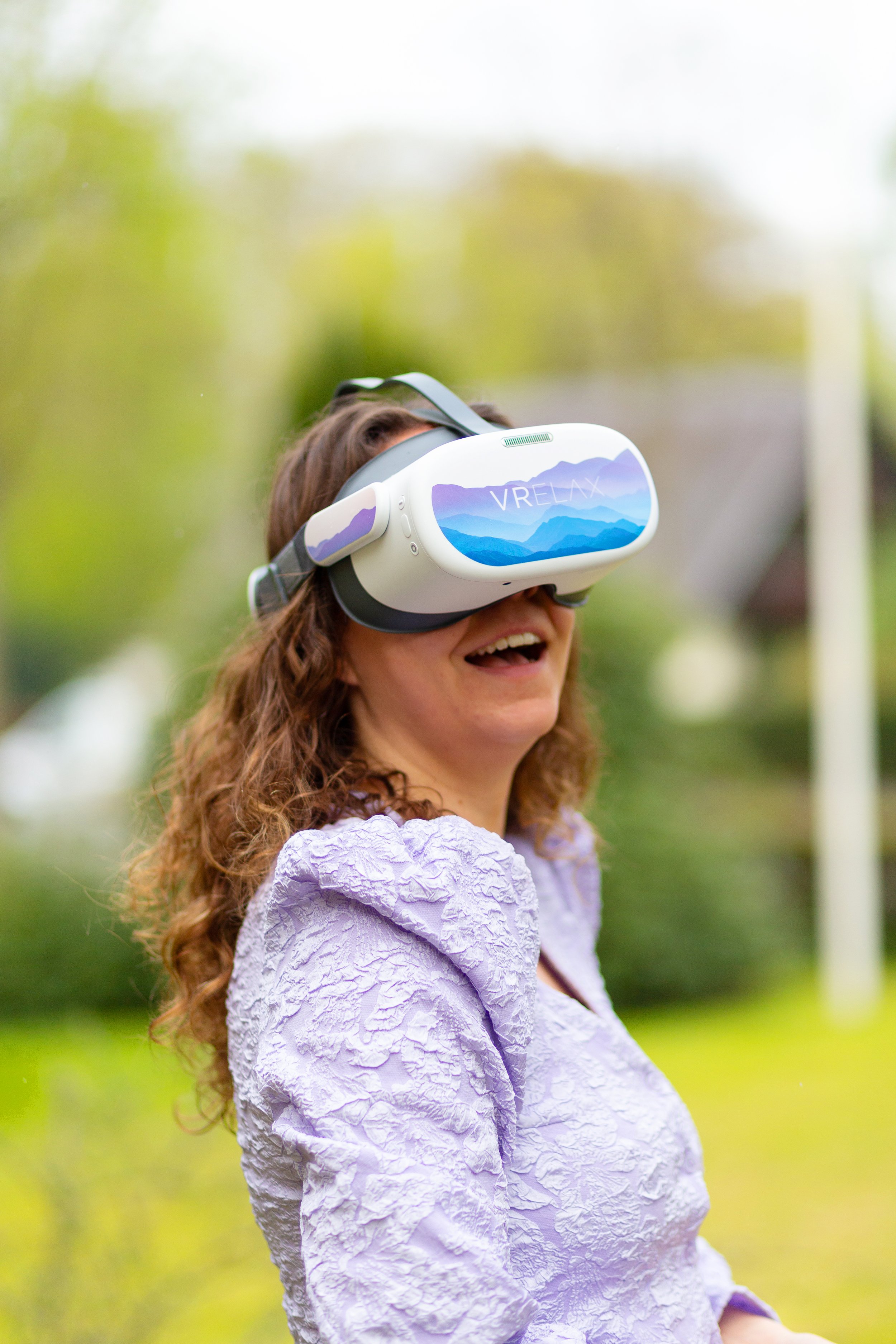 A woman smiling outdoors wearing a virtual reality headset labeled 'VRELAK' with a mountain landscape design, surrounded by greenery.