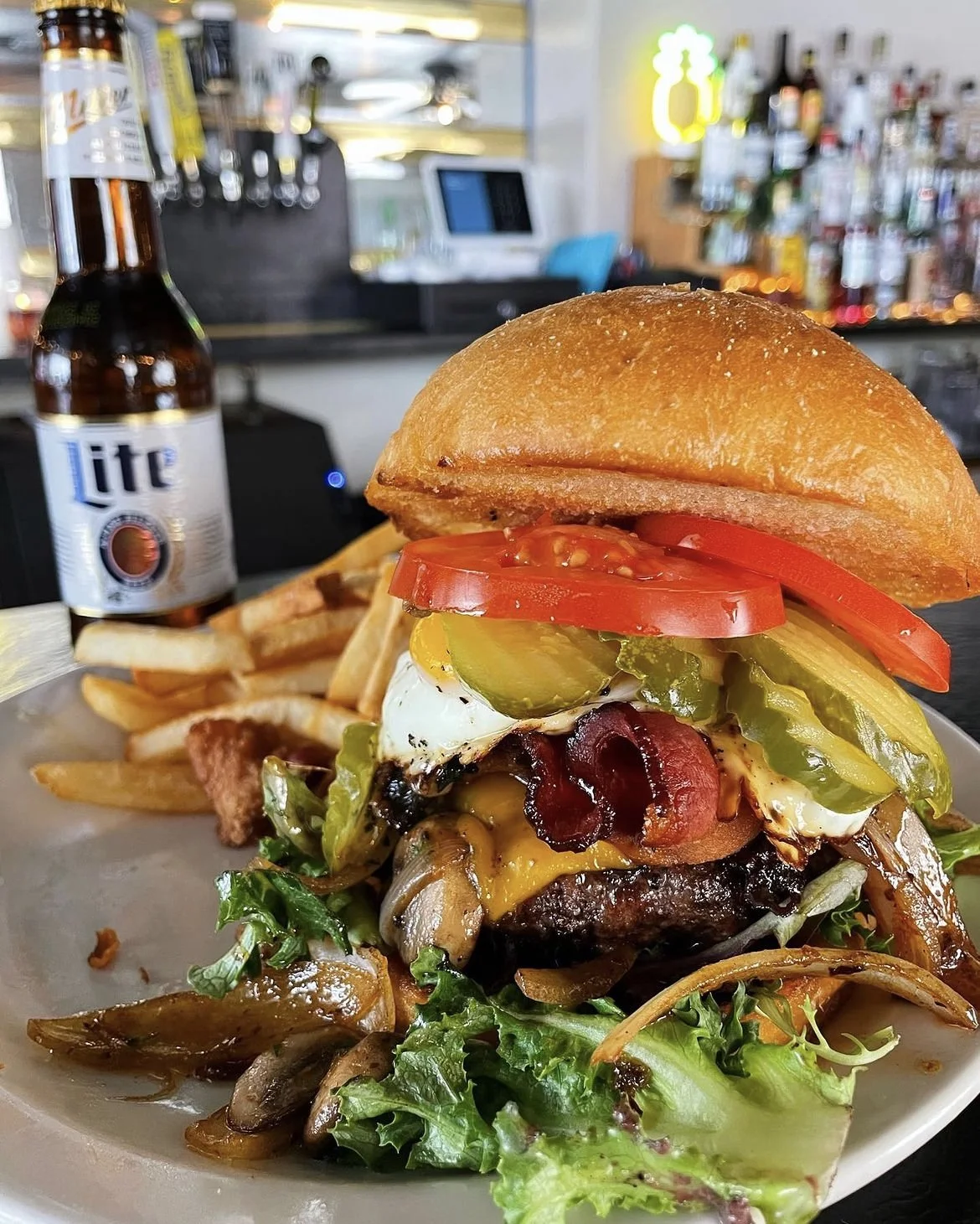 Gourmet burger with bacon, cheese, fried egg, pickles, tomatoes, and lettuce, served with fries. Bottle of Lite beer in the background on a bar counter.