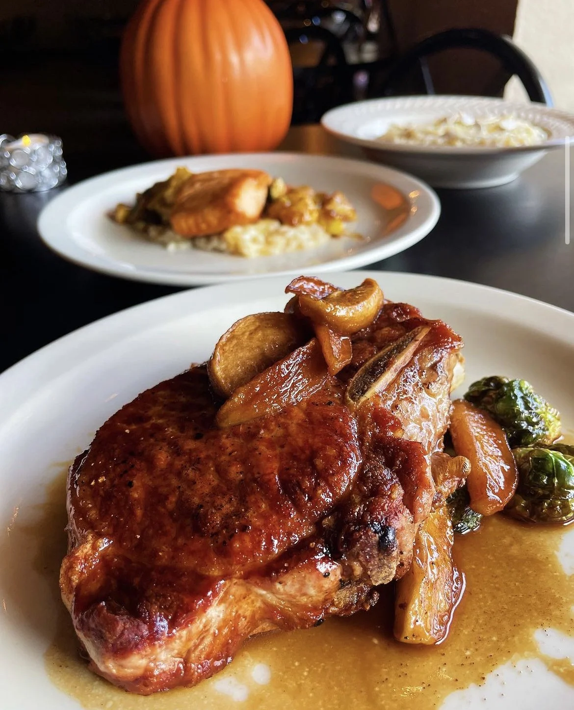 Close-up of a glazed pork chop with sautéed vegetables on a white plate. In the background, there is another plate with salmon and risotto, and a large pumpkin decor.
