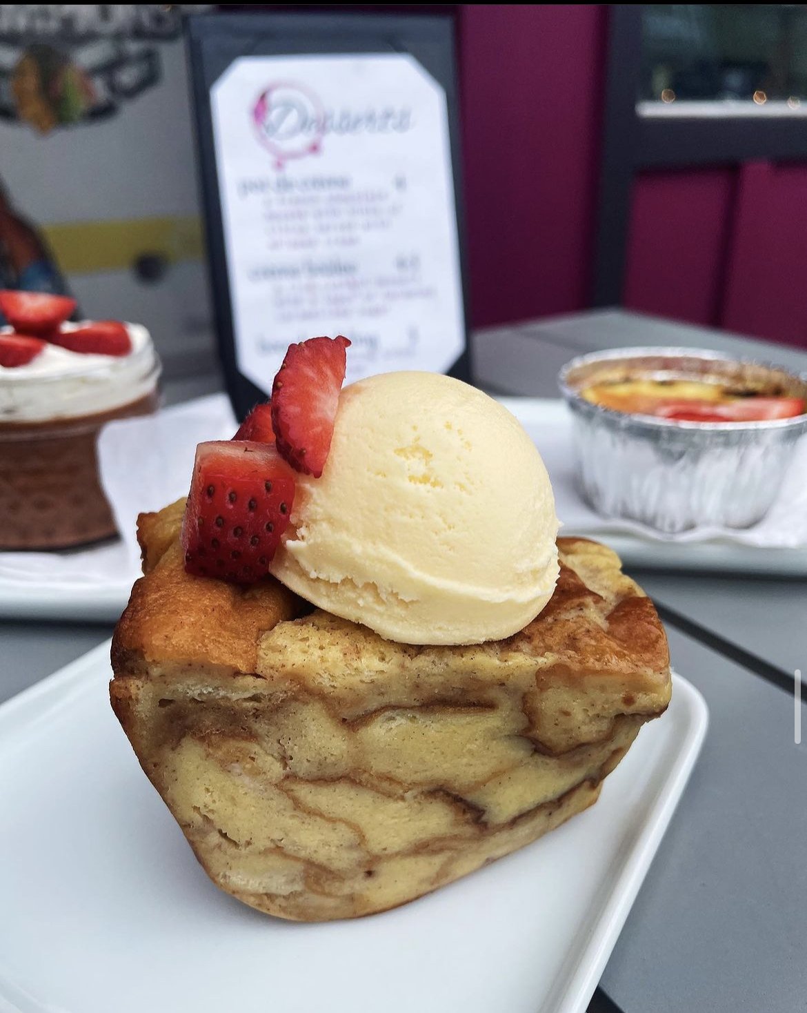 Gourmet dessert with a bread pudding topped with a scoop of vanilla ice cream and sliced strawberries, presented on a white plate; a blurred menu and additional desserts in the background.