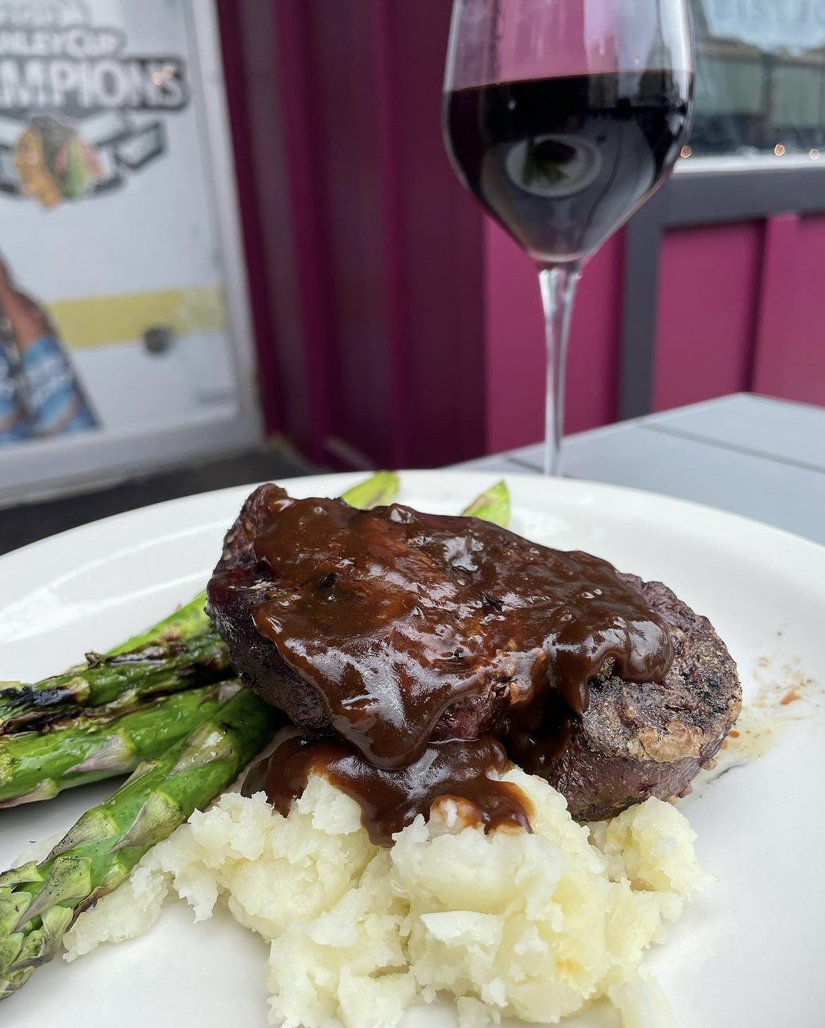 Grilled steak with brown sauce served on mashed potatoes and asparagus, accompanied by a glass of red wine.