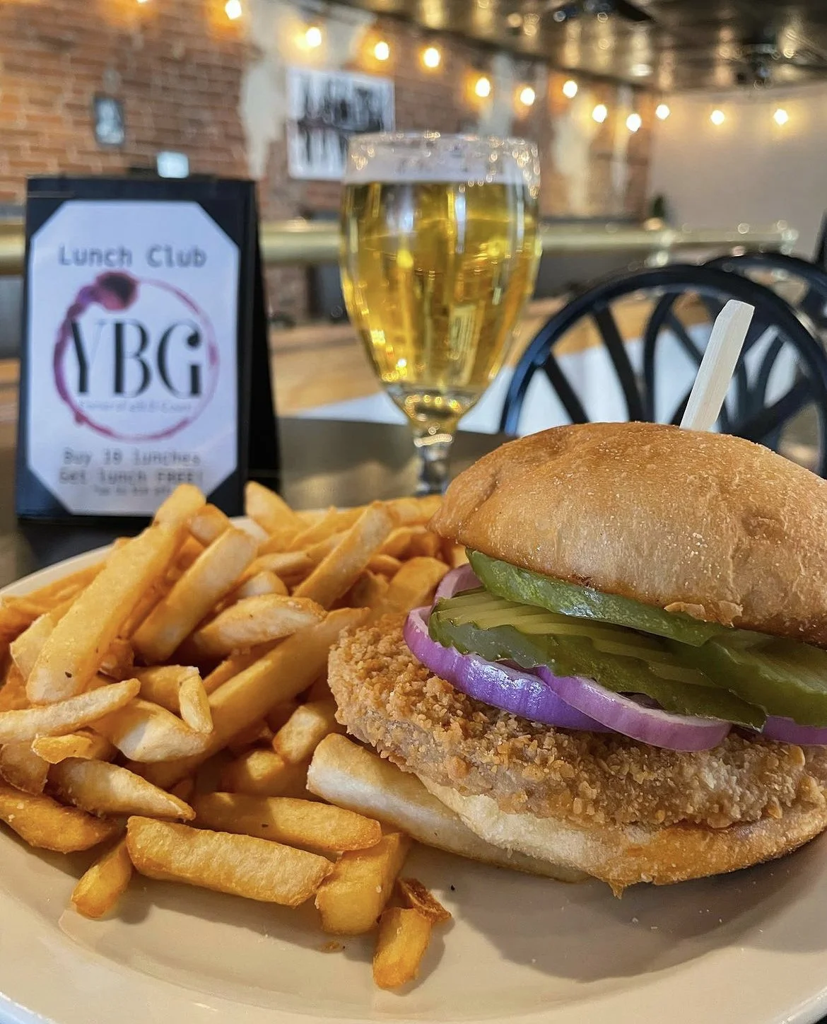 Chicken sandwich with pickles and red onion, served with fries on a plate, next to a glass of beer and a restaurant's lunch club sign on a table.