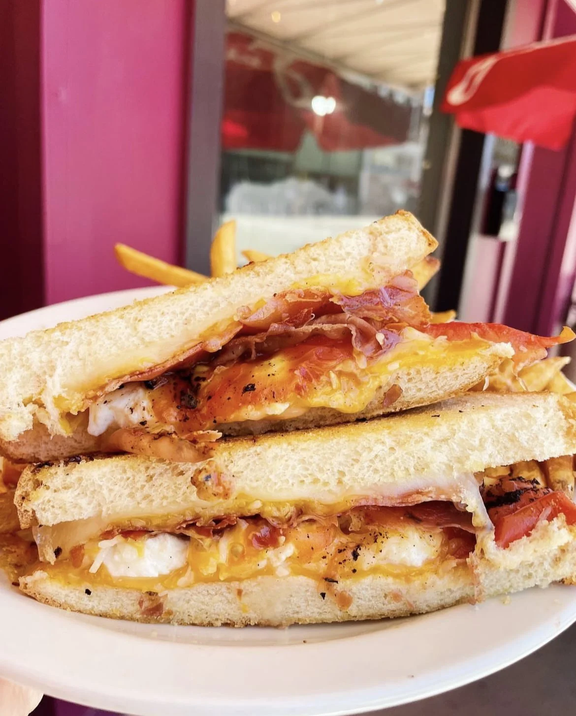 A grilled cheese sandwich with bacon and tomatoes cut in half on a white plate with fries in the background.