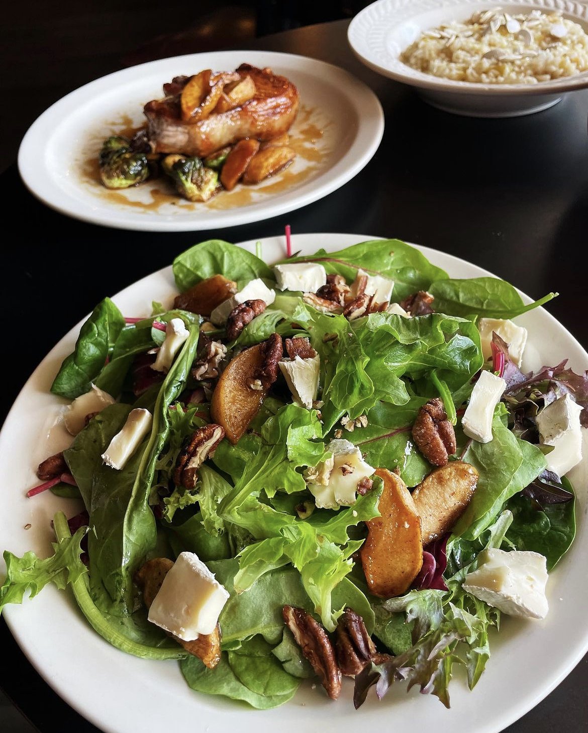Plate of mixed green salad with brie cheese, candied nuts, and apple slices; background showing pork with Brussels sprouts and risotto.