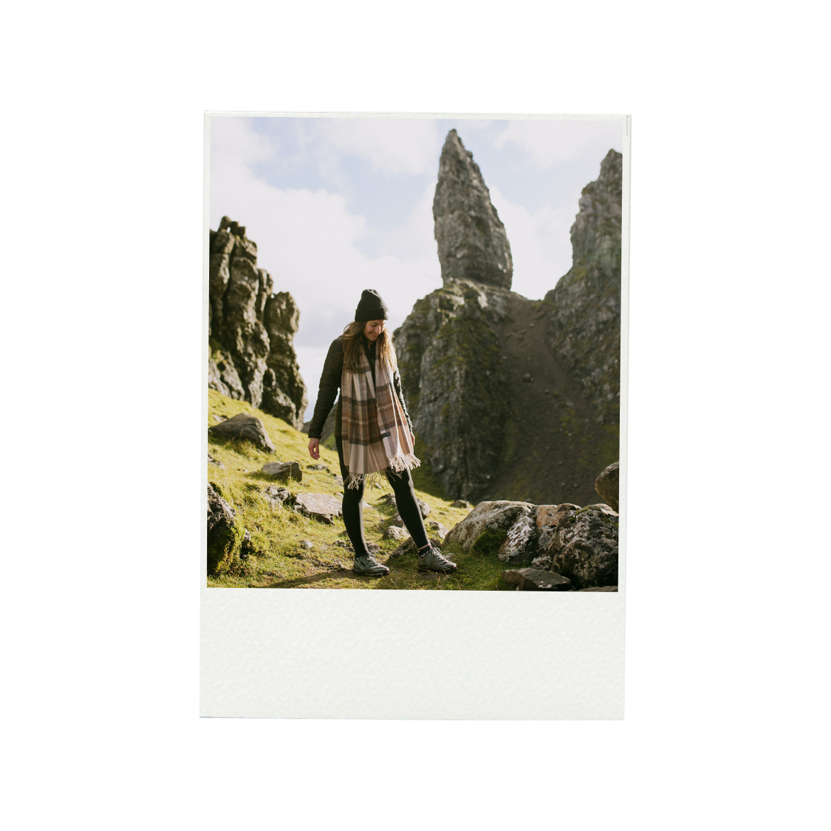 A young woman in outdoor clothing, including a black beanie, hiking boots, a scarf, and a jacket, walking on a grassy trail with rocky terrain. Behind her are two large, pointed rock formations under a partly cloudy sky.