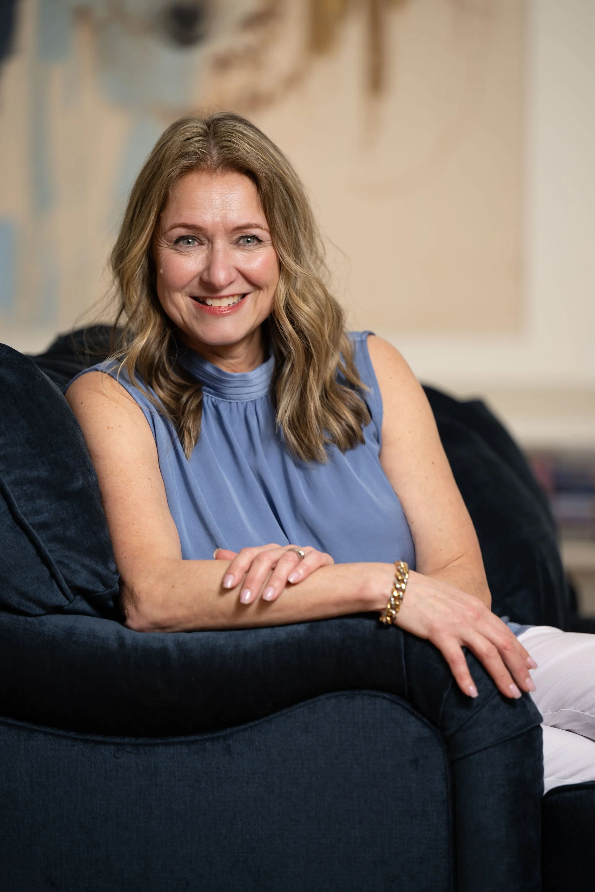 Mandy Baker, A smiling woman with wavy blonde hair, wearing a blue blouse, sitting on a black couch ready to help you with your finances and wealth.