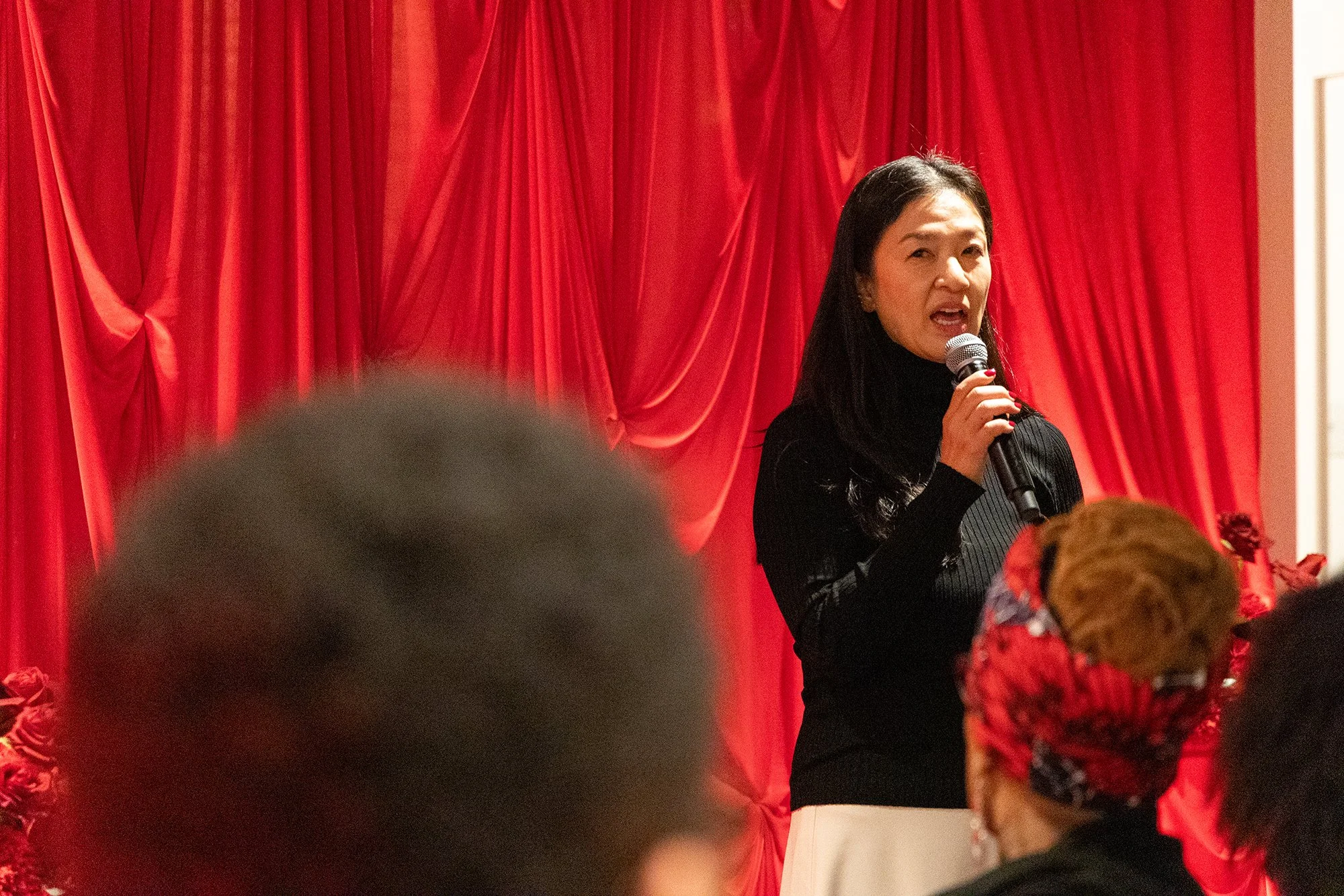 Woman speaking into a microphone with a red curtain backdrop, audience in foreground.