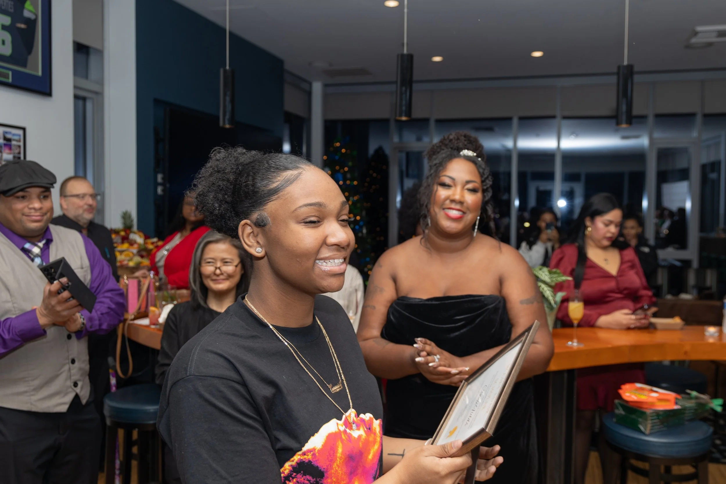A group of people at a celebration or party, with two women in the foreground smiling, one holding a framed certificate or picture, and others in the background. The setting appears to be indoors with festive decorations and drinks.