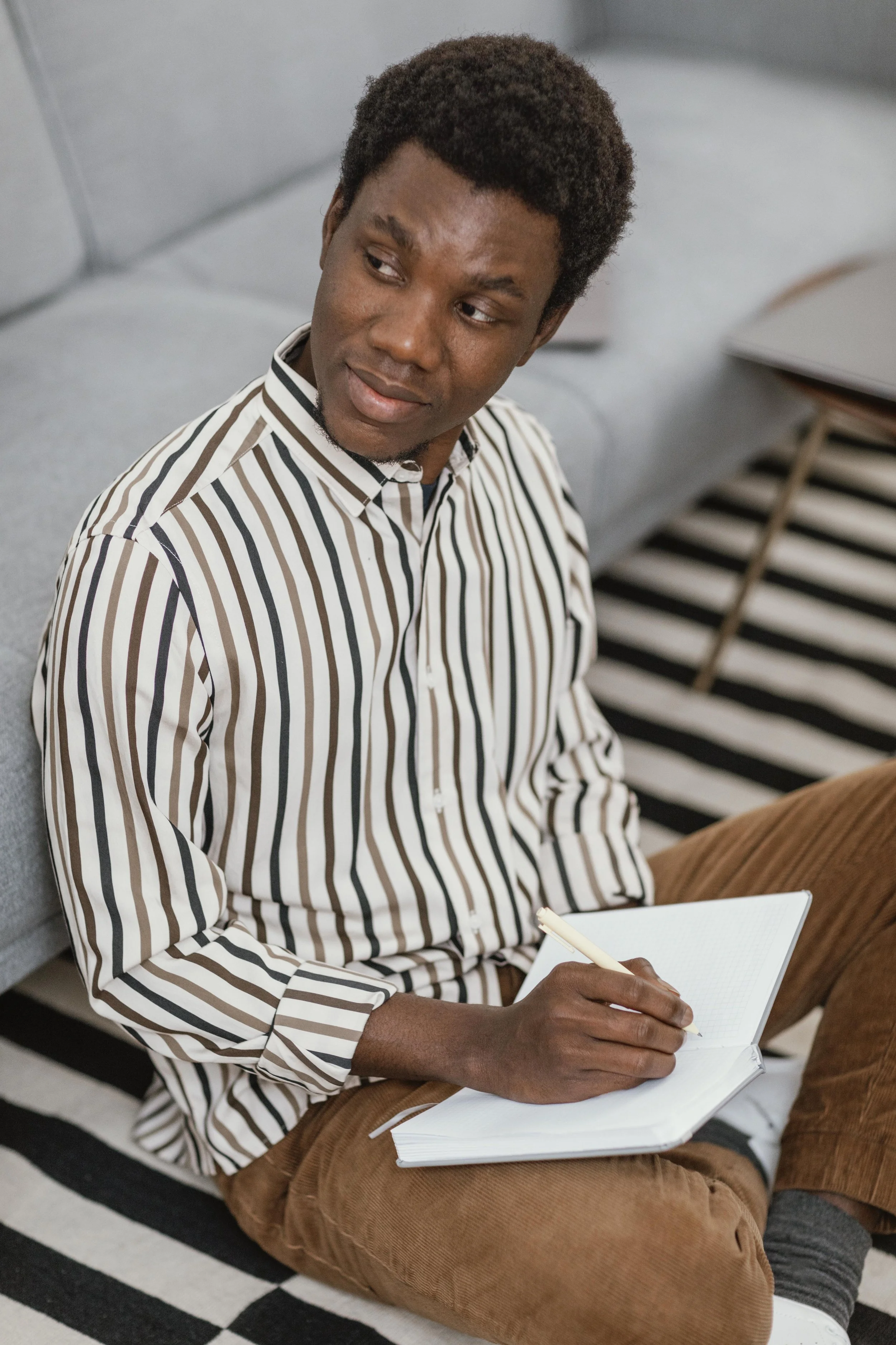A man sitting on the floor, writing in a notebook, with a gray sofa and a black and white striped rug in the background.