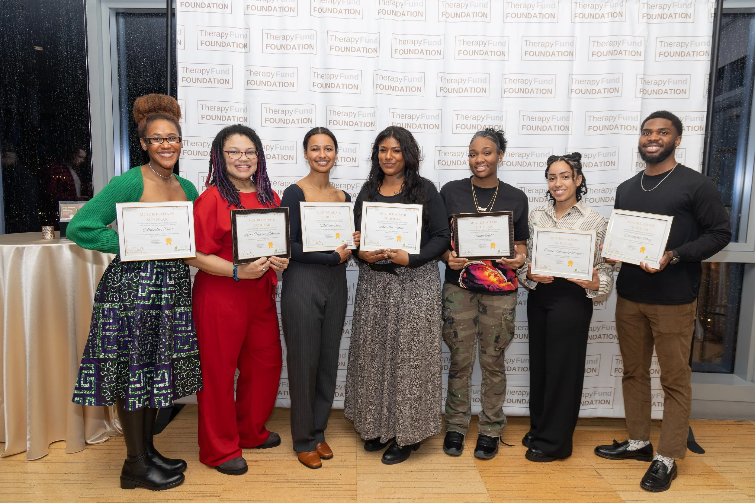 Group of seven diverse young adults holding certificates and smiling at an awards event, standing in front of a backdrop with 'Therapy Fund Foundation' repeated on it.