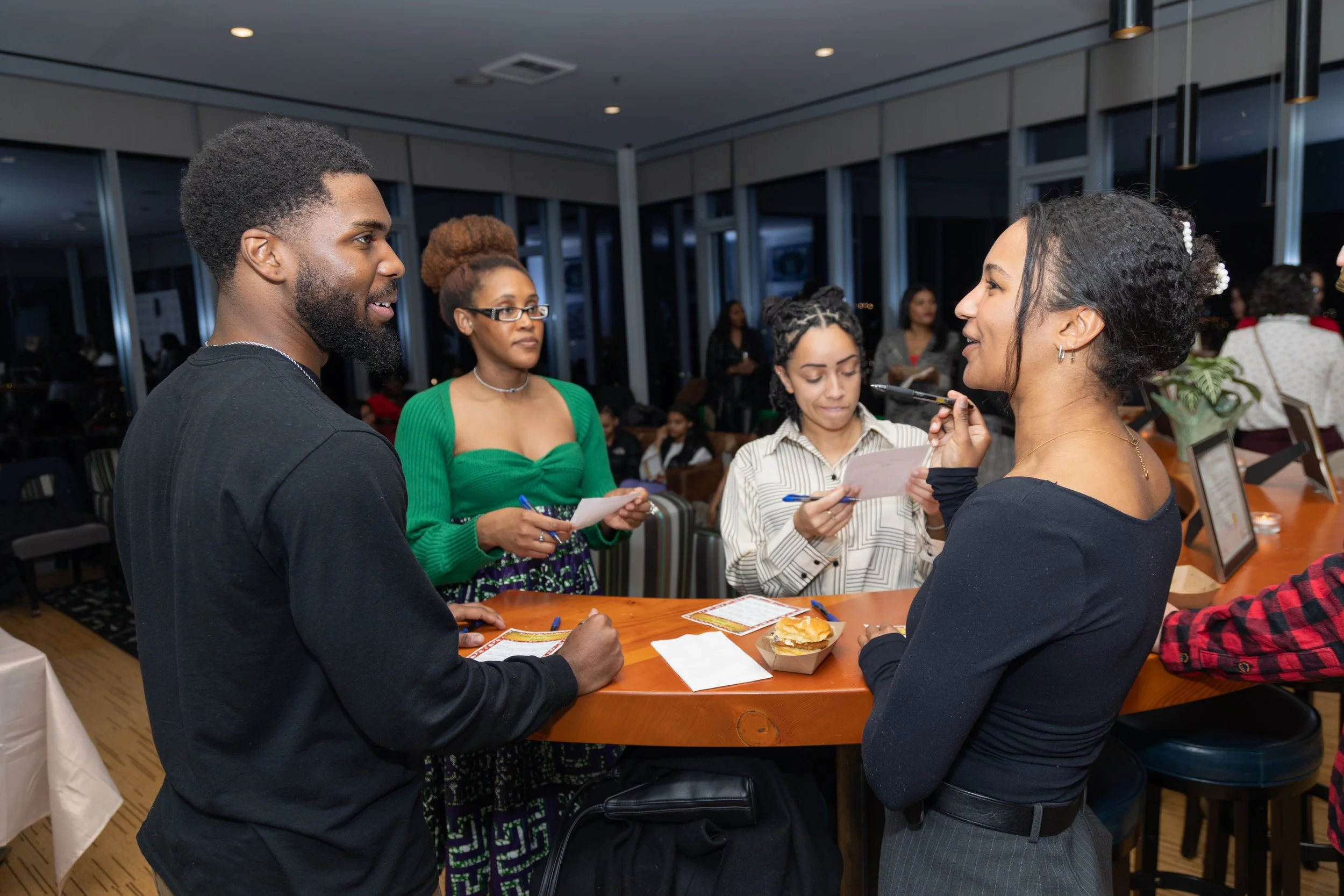 A group of five people gathered around a bar in a restaurant, engaged in conversation. One woman with curly hair is holding a pen and a notepad, another woman with glasses and a green top is writing, a man with a beard in a black shirt is smiling, an