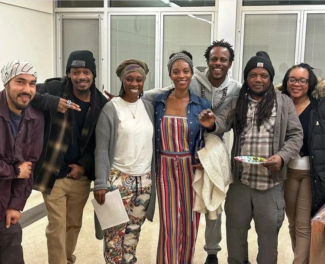 A group of eight diverse people standing together indoors, smiling, with some holding papers or plates, in front of large windows.