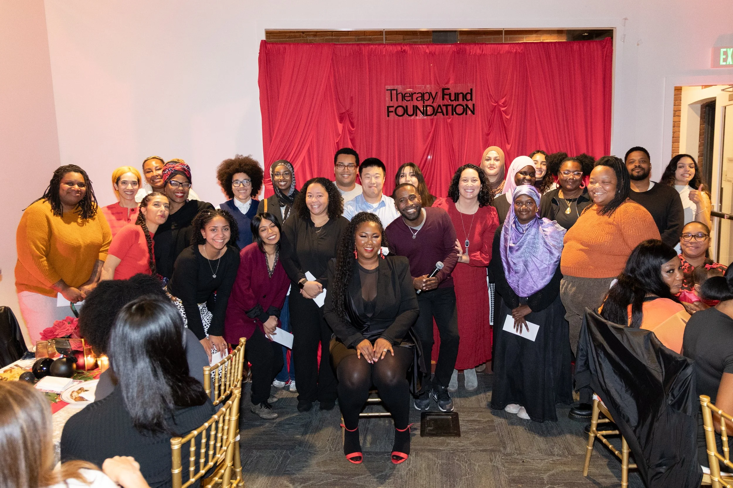 Group of diverse people gathered for a photo at an event, standing in front of a red curtain with a sign that says 'Therapy Fund FOUNDATIONS', with some seated at tables and others standing, smiling at the camera.