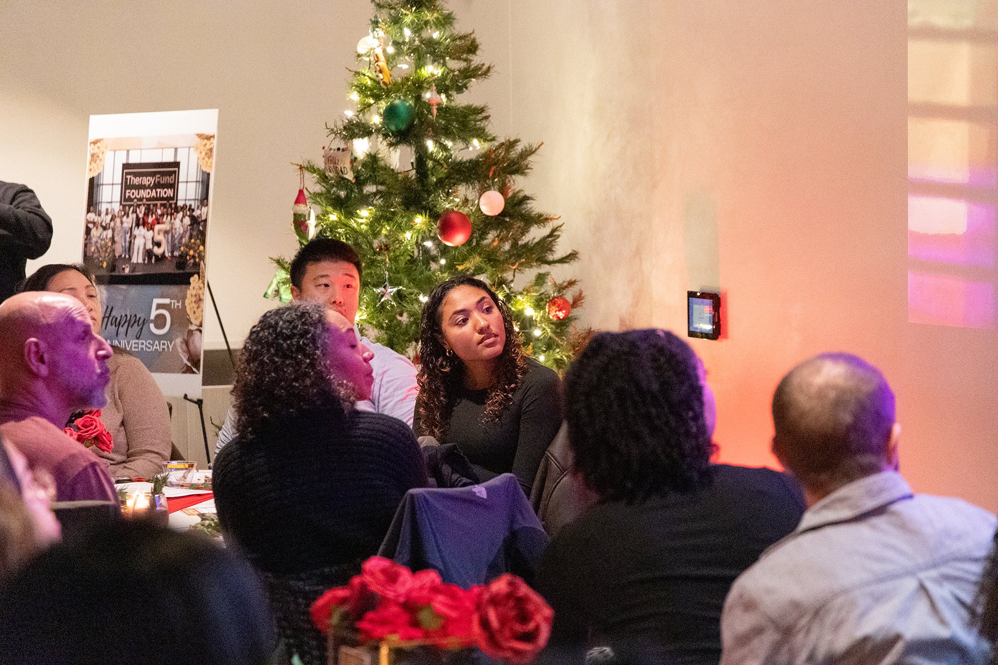 People sitting around a table listening to a presentation or performance, with a decorated Christmas tree nearby at a holiday gathering.