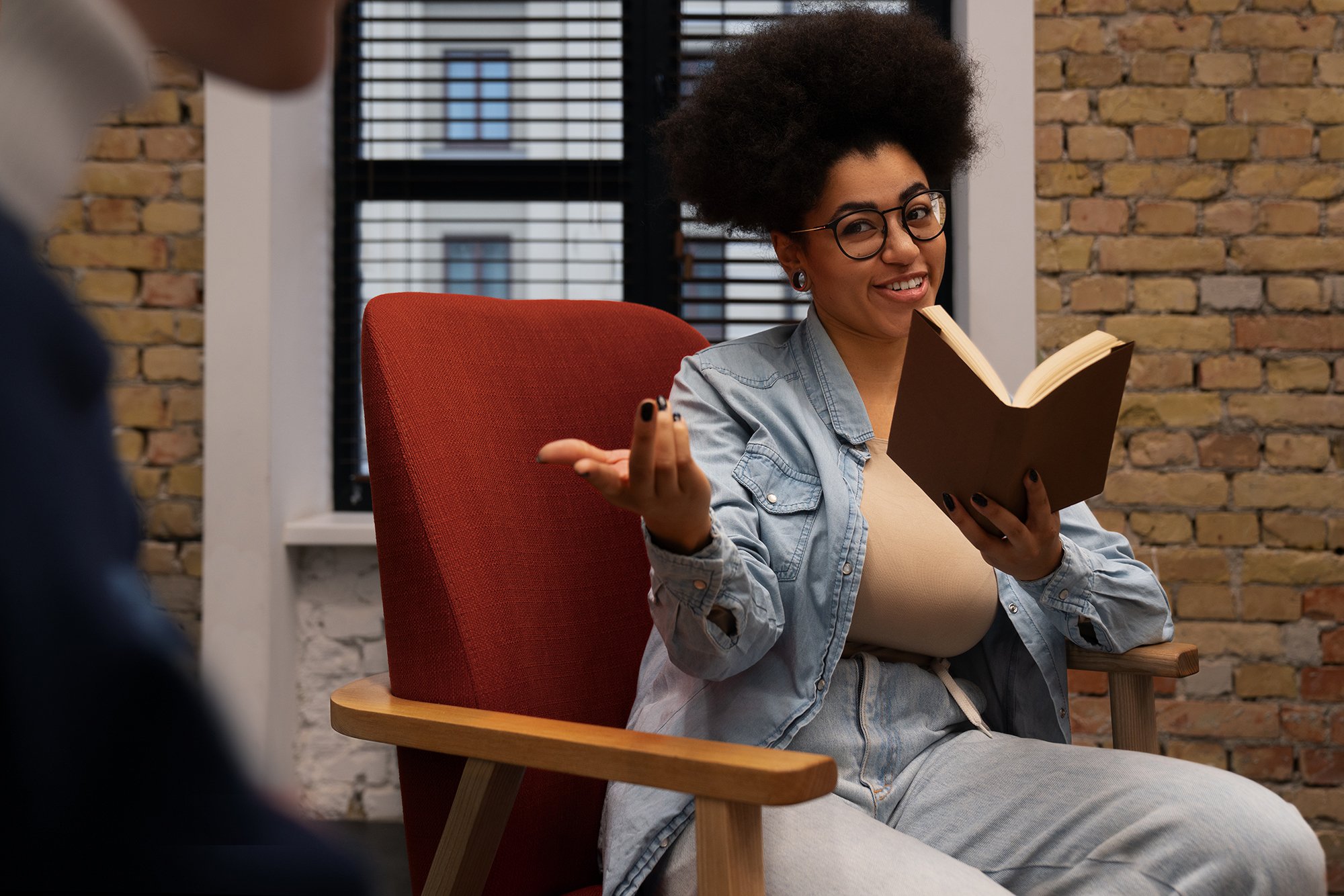 A woman with curly hair, glasses, and earrings smiling and gesturing while holding an open book, sitting on a red chair in front of a brick wall and window blinds.