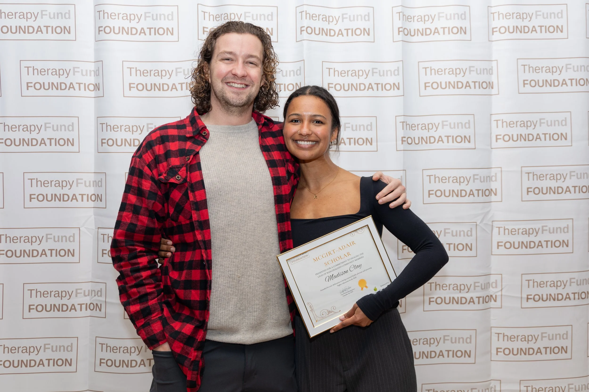 A woman holding a scholarship certificate standing next to a man at a Therapy Fund Foundation event, both smiling for the camera in front of a backdrop with the foundation's name.