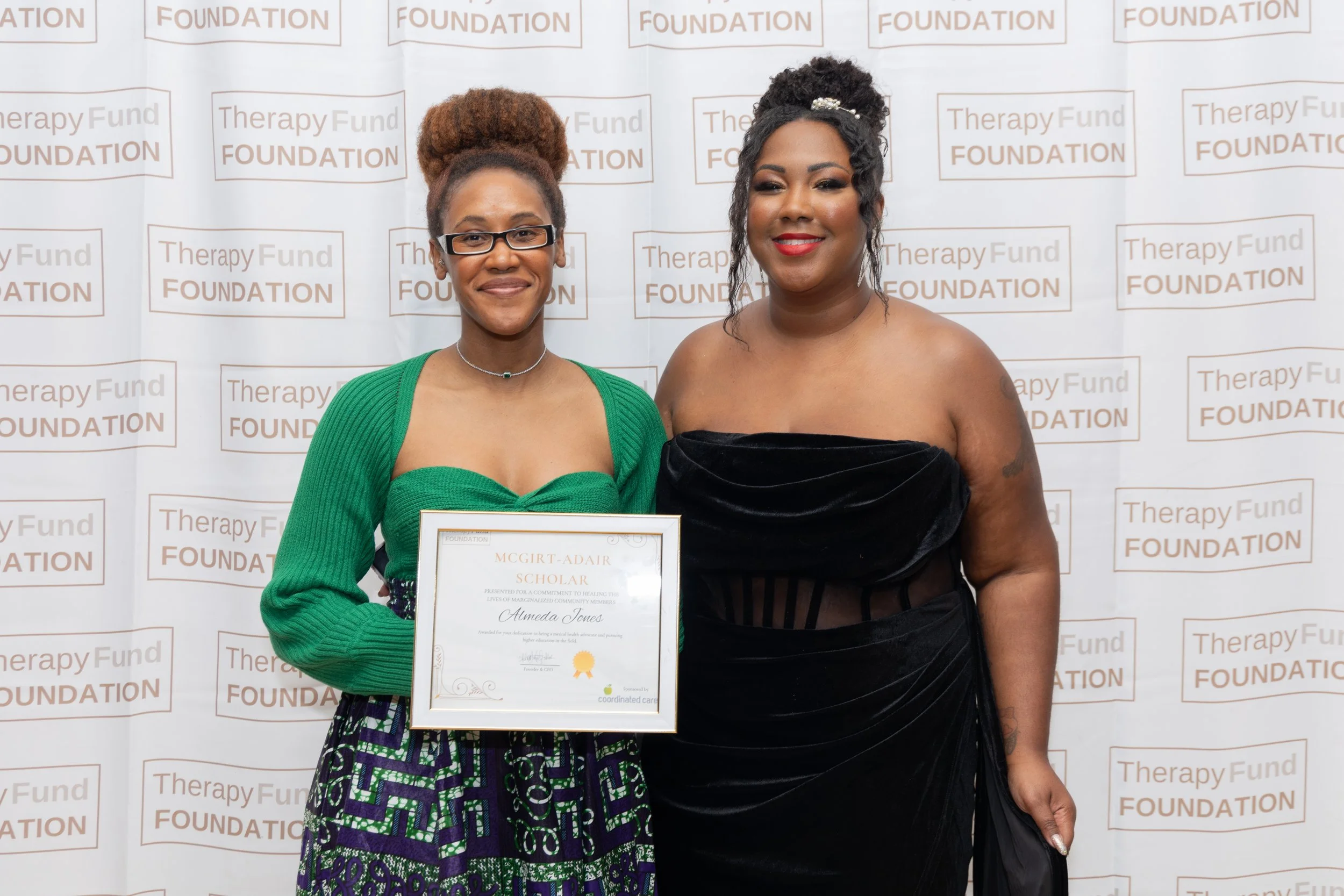 Two women standing together in front of a backdrop with the logo of the Therapy Fund Foundation. The woman on the left, with glasses and her hair in a bun, is holding a framed certificate and wearing a green top and a patterned skirt. The woman on th