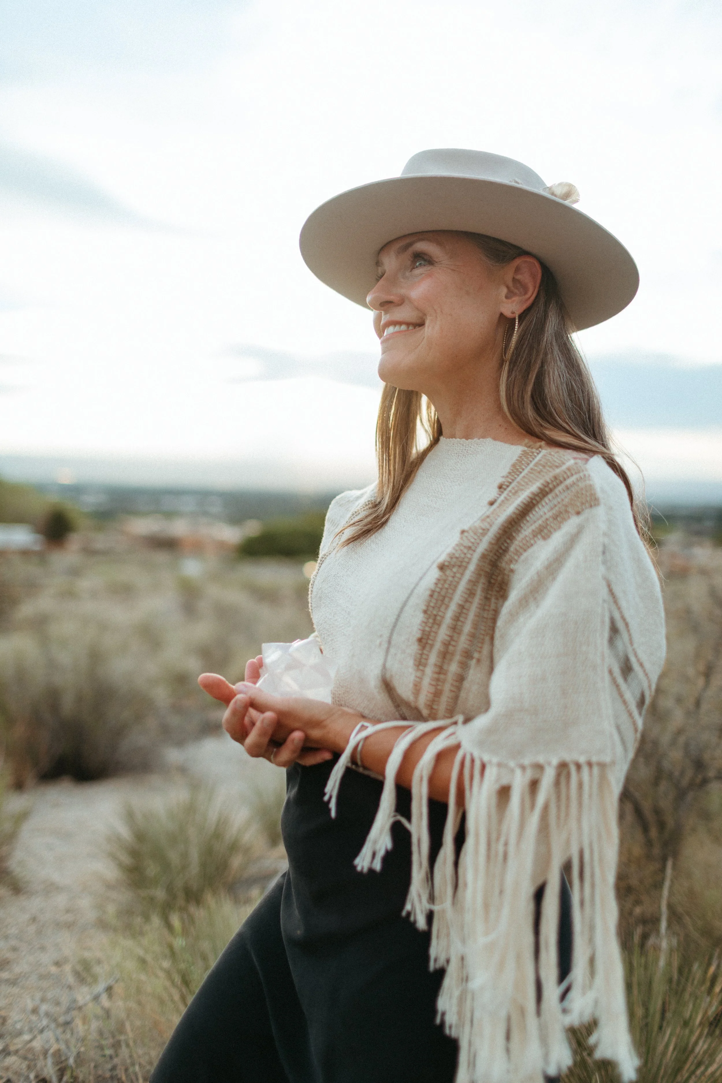 A woman with light brown hair, wearing a large beige hat with a decorative element, a cream-colored sweater with brown stripes, and black pants, standing outdoors in a natural landscape with tall grasses and a cloudy sky.