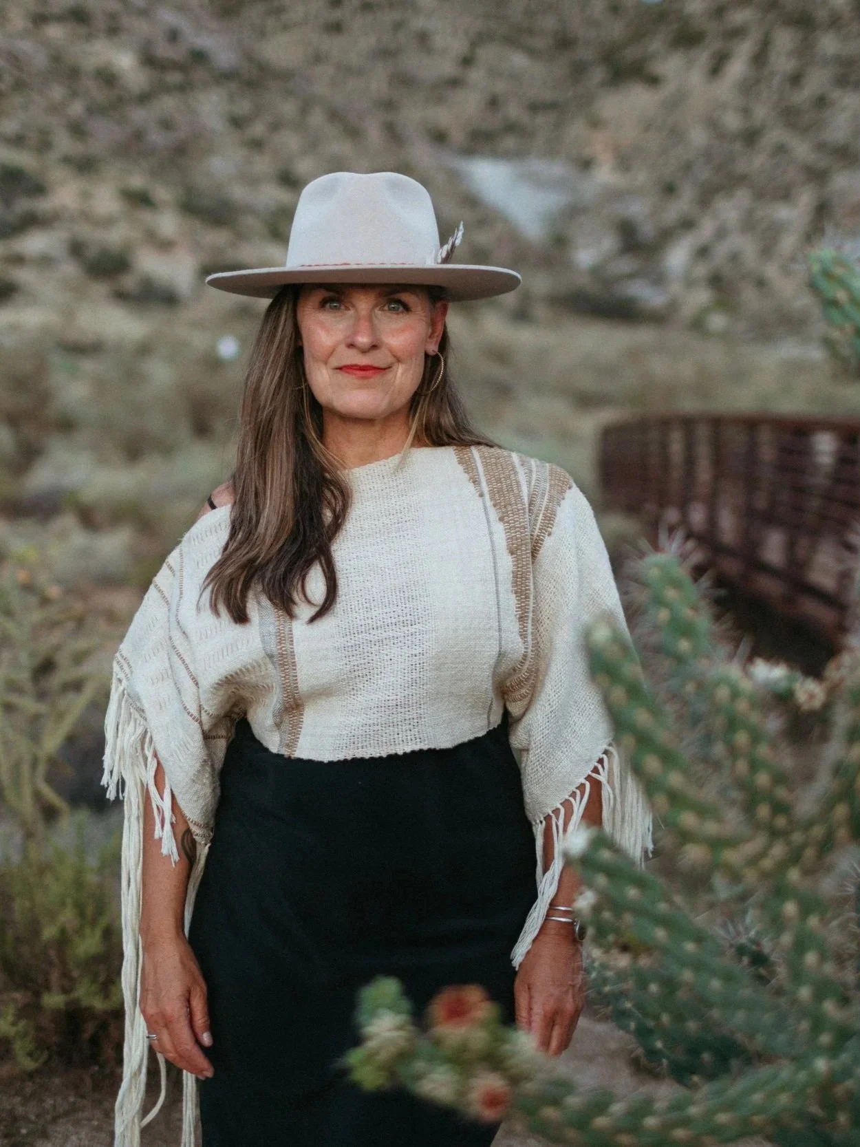 A woman standing outdoors in a desert landscape with rocks and cacti. She is wearing a large gray hat, a cream-colored shawl, a black skirt, and a bracelet.