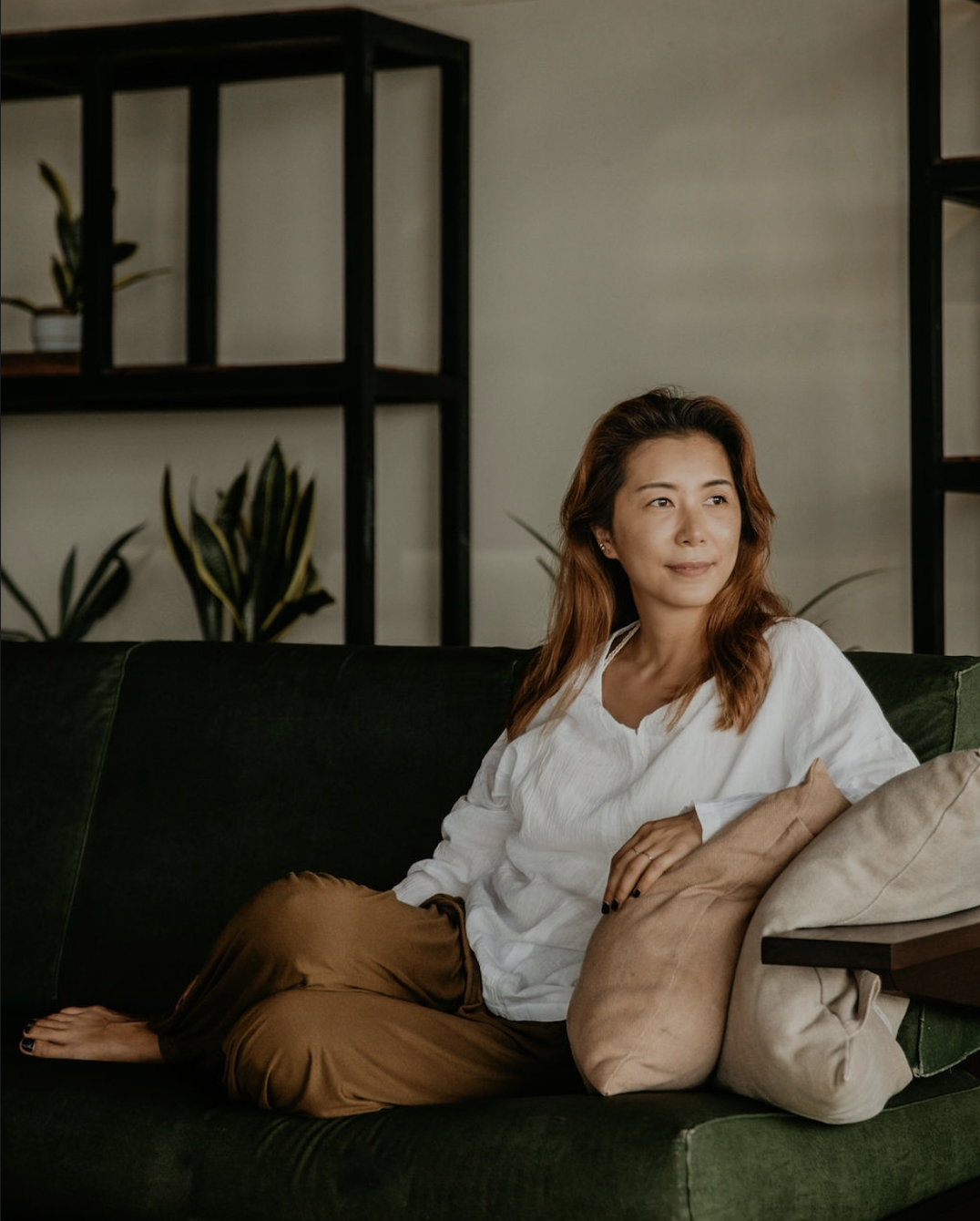 A woman with wavy brown hair sitting on a dark green sofa, leaning on beige pillows, in a living room with shelves and potted plants in the background.
