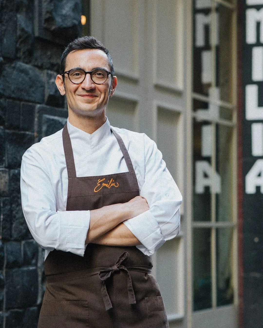 Dino the head chef and owner of Trattoria Emilia smiles outside the restaurant in a white chef shirt and dark apron with "Emilia" embroidered on the chest. A black brick wall and blurred doorway frame the portrait.