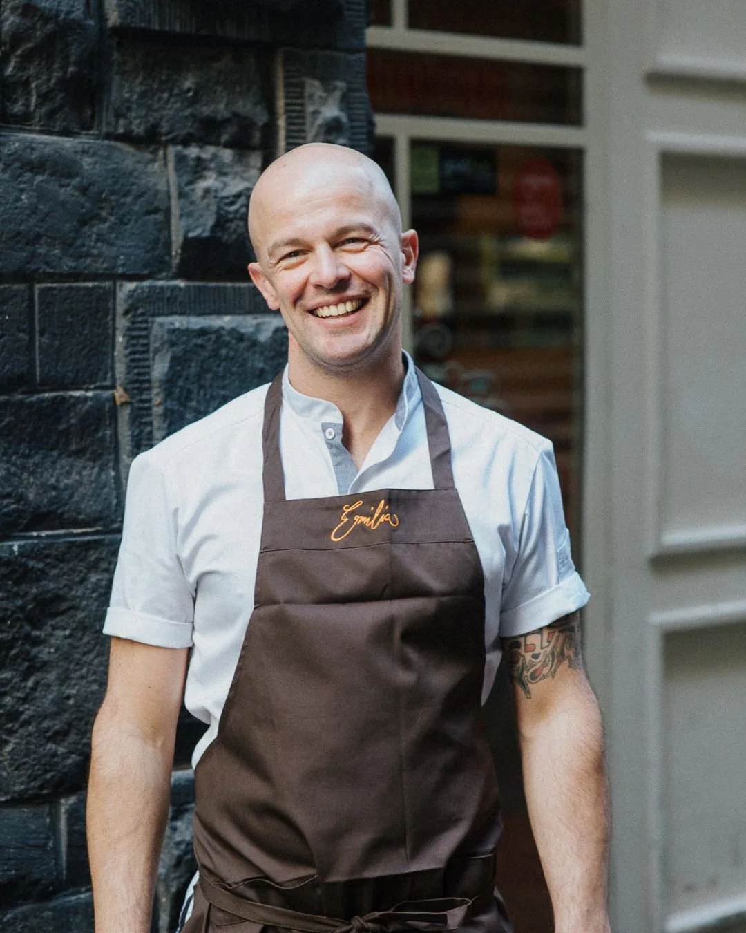 Francesco the head chef and owner of Trattoria Emilia smiles outside the restaurant in a white chef shirt and dark apron with "Emilia" embroidered on the chest. A black brick wall and blurred doorway frame the portrait.