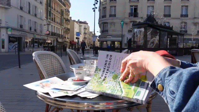 Outdoor café table on a city street with newspapers, coffee cups, and a person reading a booklet about environmentalism.