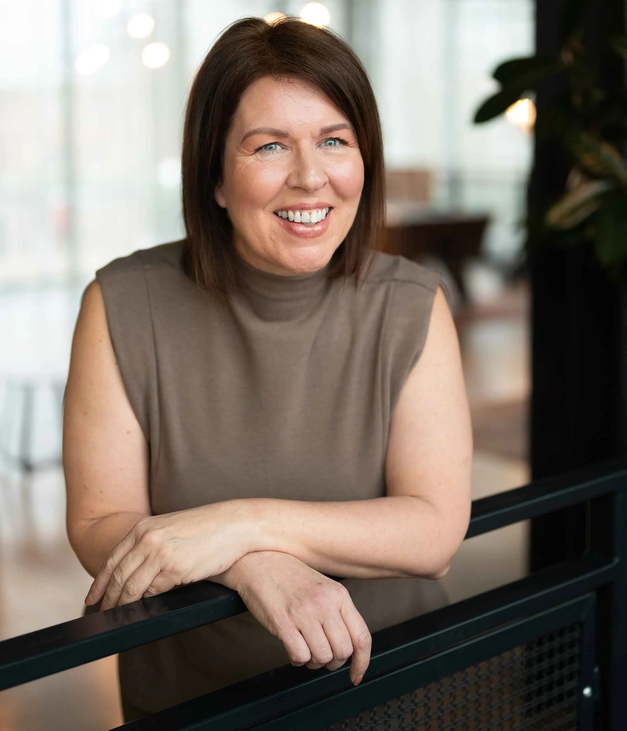 A woman with shoulder-length brown hair, smiling, wearing a sleeveless taupe top, leaning on a black railing in a well-lit indoor setting.