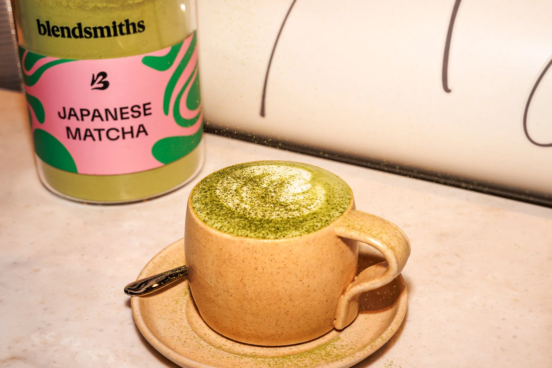 A beige ceramic cup filled with matcha latte topped with matcha powder, placed on a matching saucer with a spoon. Behind it is a container labeled 'Japanese Matcha' with green and pink design, and a yellow lid labeled 'blendsmiths'.