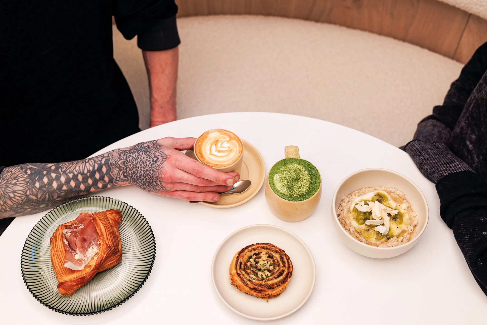 A white table with a pastry, two drinks, and a bowl of rice pudding with toppings. Two people are sitting at the table, one with a tattooed arm reaching for a latte with latte art, and the other with a dark sleeve.