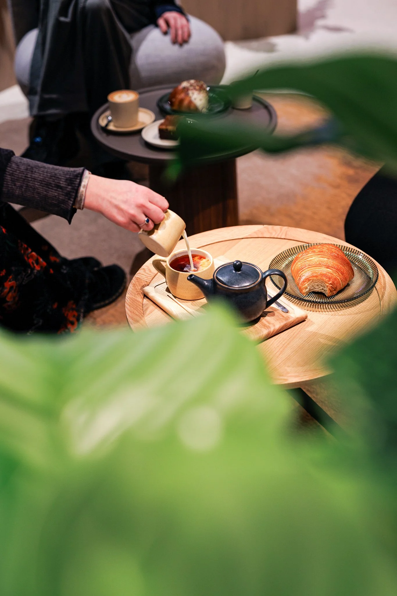 Person pouring cream into a cup of tea on a wooden tray with a croissant beside it. In the background, a table with a cappuccino, a plate with a pastry, and another plate with a cake are visible.