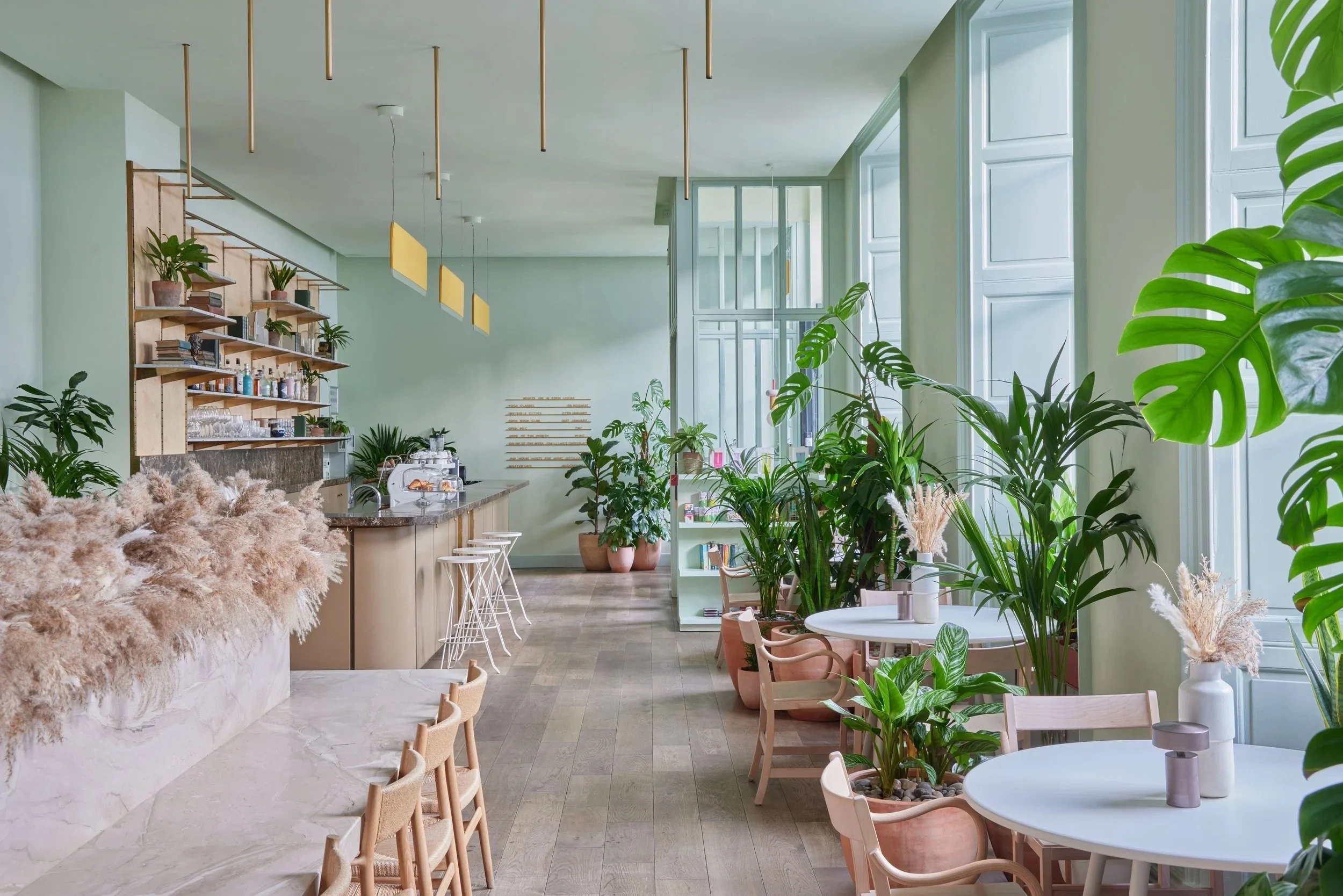 Bright green interior of a modern cafe or restaurant with large windows, green plants, and cozy seating areas with pink and white furniture.