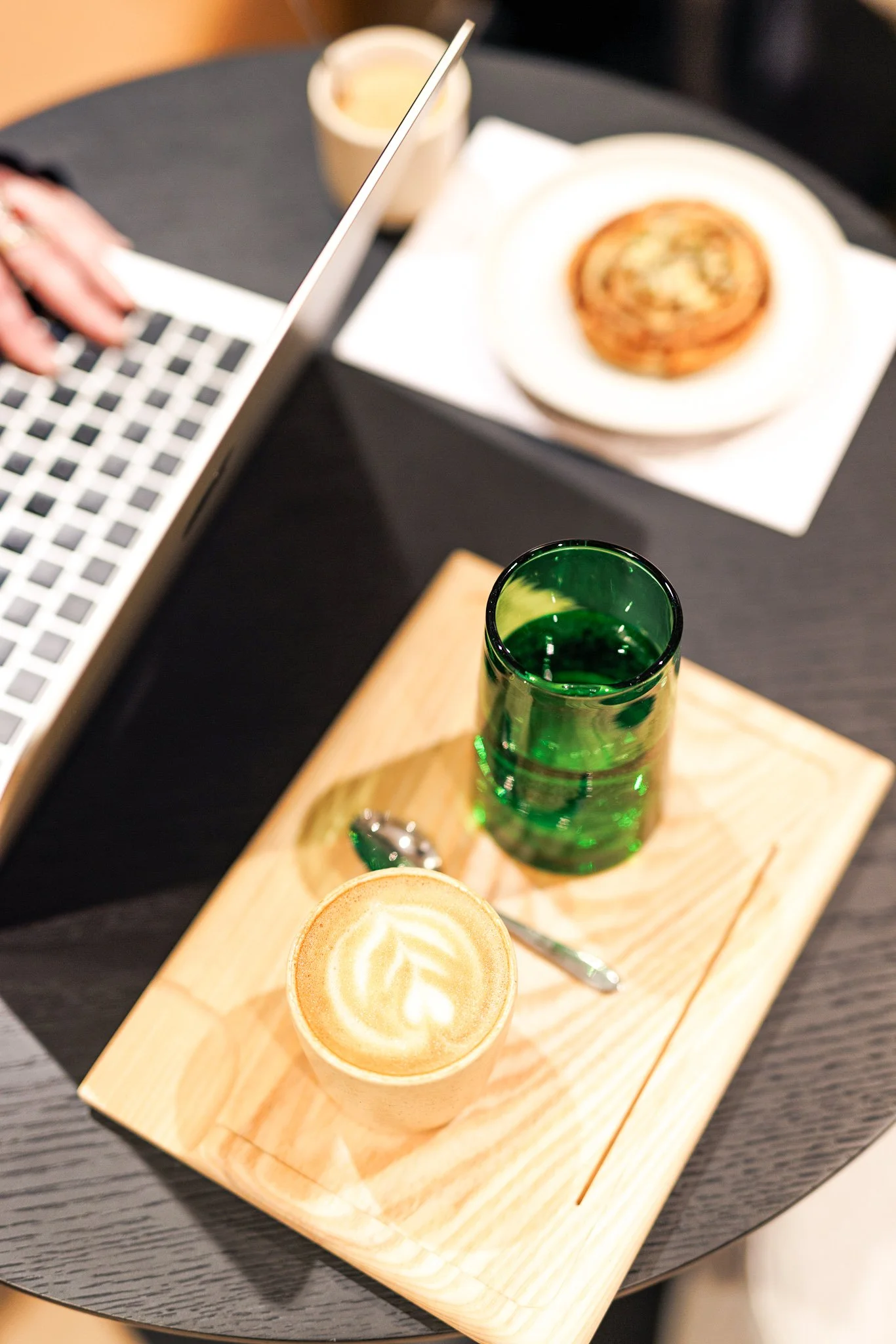 A wooden tray holding a green glass, a small cup of latte with latte art, and a spoon. In the background, a plate with a pastry and a cup of coffee are on a dark table, with a laptop partially visible.