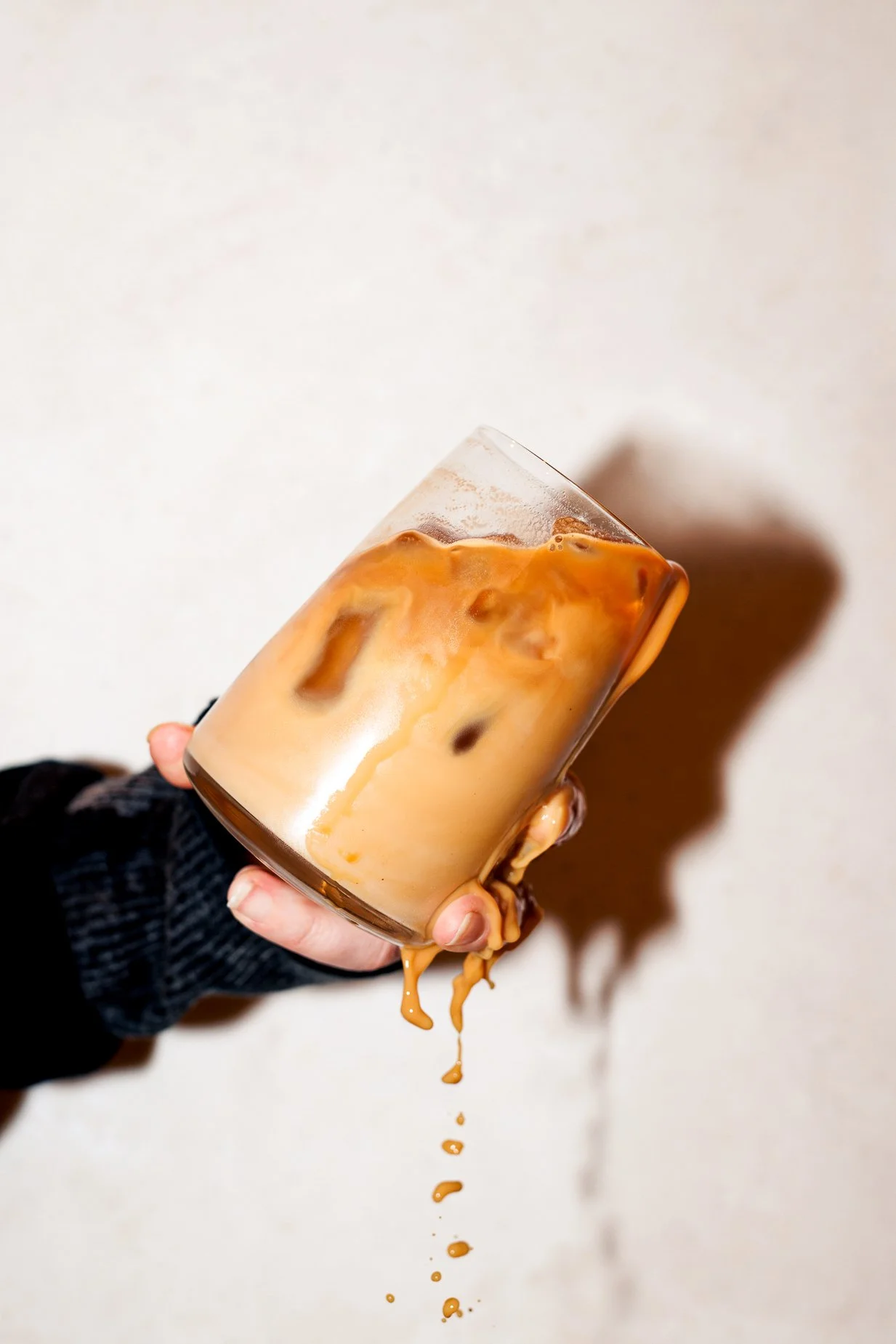 A hand holding a glass of iced coffee with milk, spilling over the side, against a plain background.
