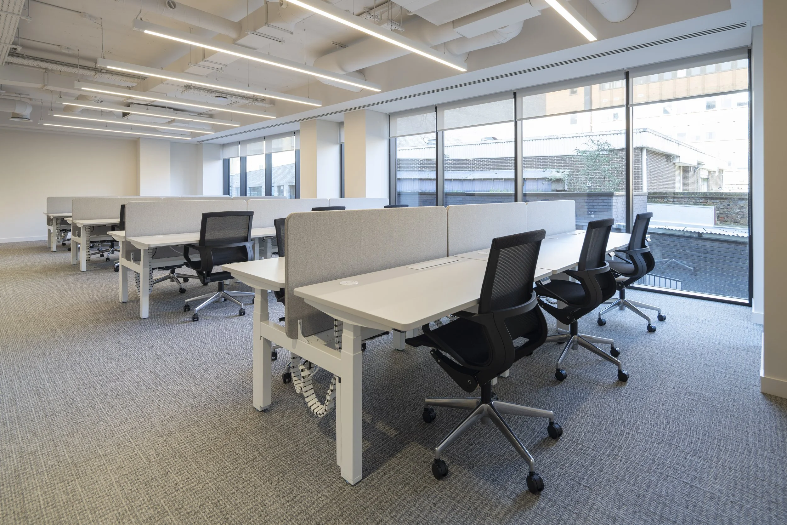 Modern office space with white desks and black office chairs arranged in rows, large windows letting in natural light, and ceiling with white industrial pipes and linear lighting