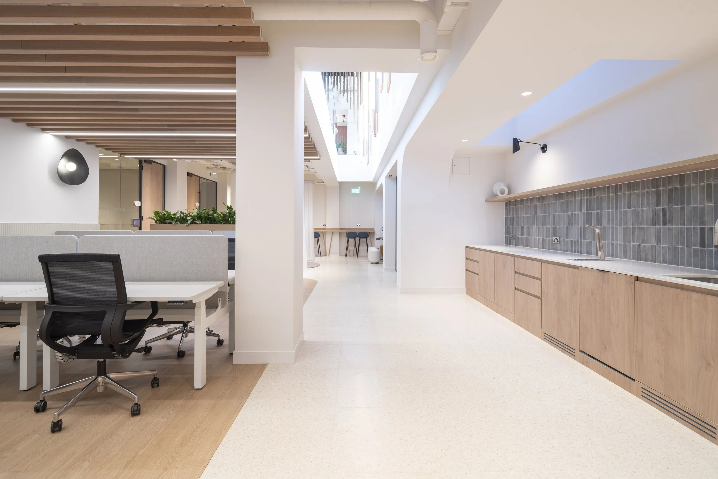 Modern office space with workspace desks and chairs, a kitchenette with wooden cabinets and a gray tile backsplash, and a bright, open layout with natural light.